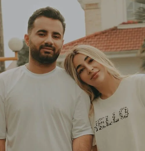A young man and woman stand close together outside, smiling softly at the camera. The man has short dark hair and a beard, wearing a white T-shirt. The woman has long blonde hair, wearing a white T-shirt with the word 'CHLOE' printed on it. There is a building with a red-tiled roof and a lamp post in the background.