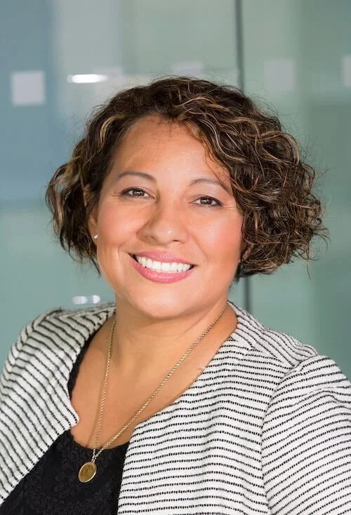 Portrait of a middle-aged woman smiling, with curly short hair, wearing a striped blazer over a black top, in an office setting.