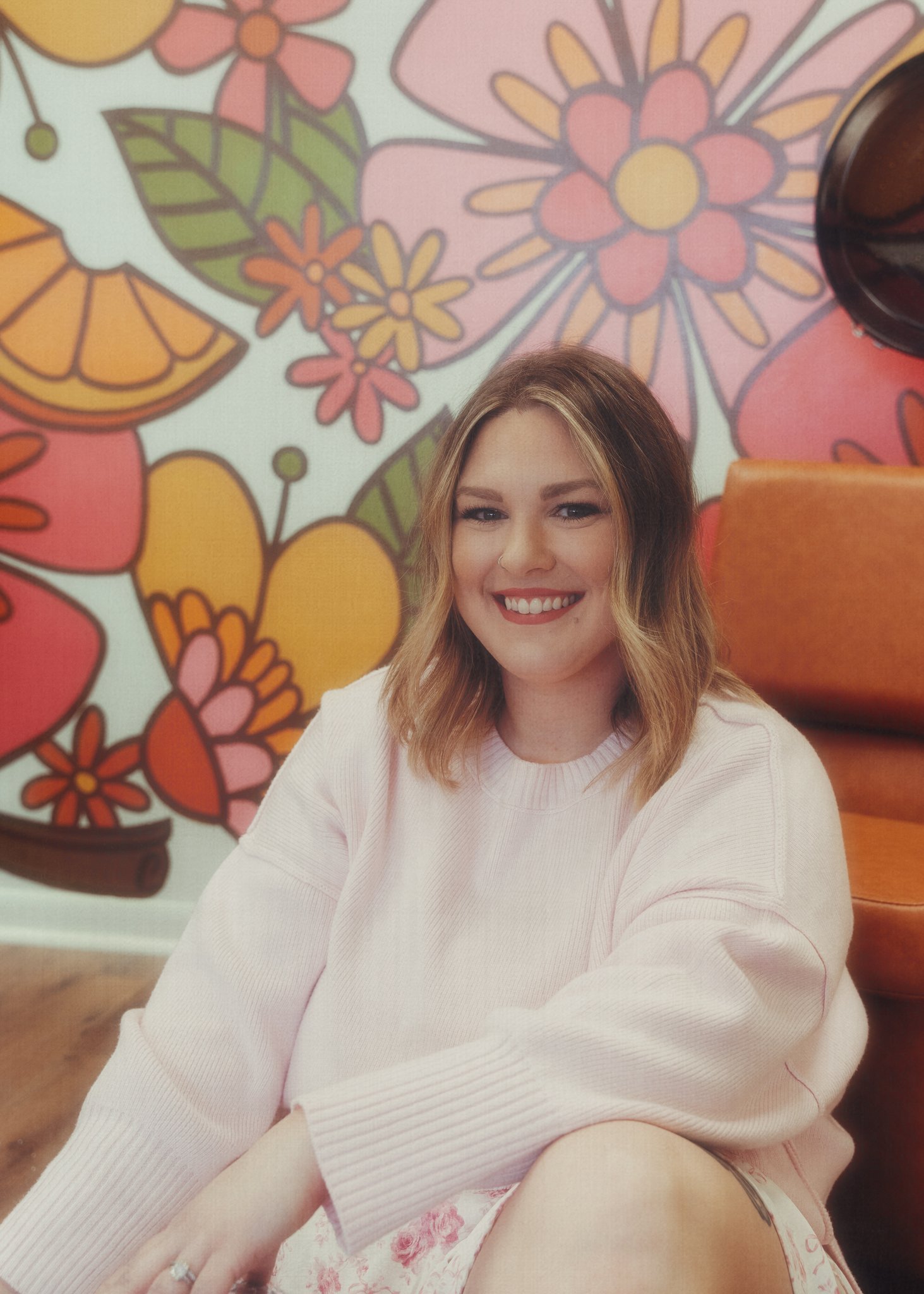 Jordan smiles in front of the flower mural. She has short blonde hair and wears a white knit sweater.