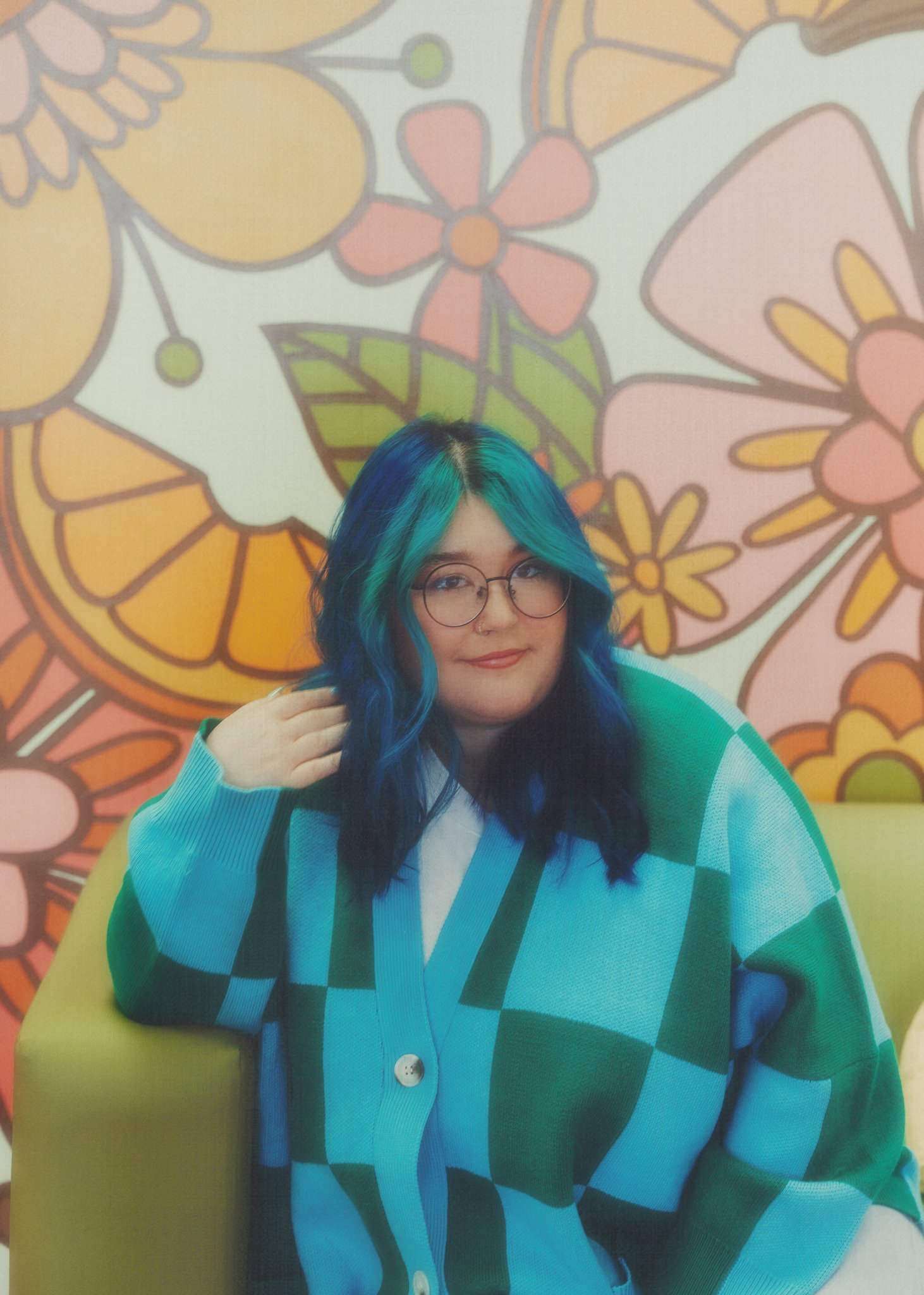 Gail poses in front of the flower mural with her hand in her teal and blue hair. She wears a teal and green checkered sweater that matches her hair.