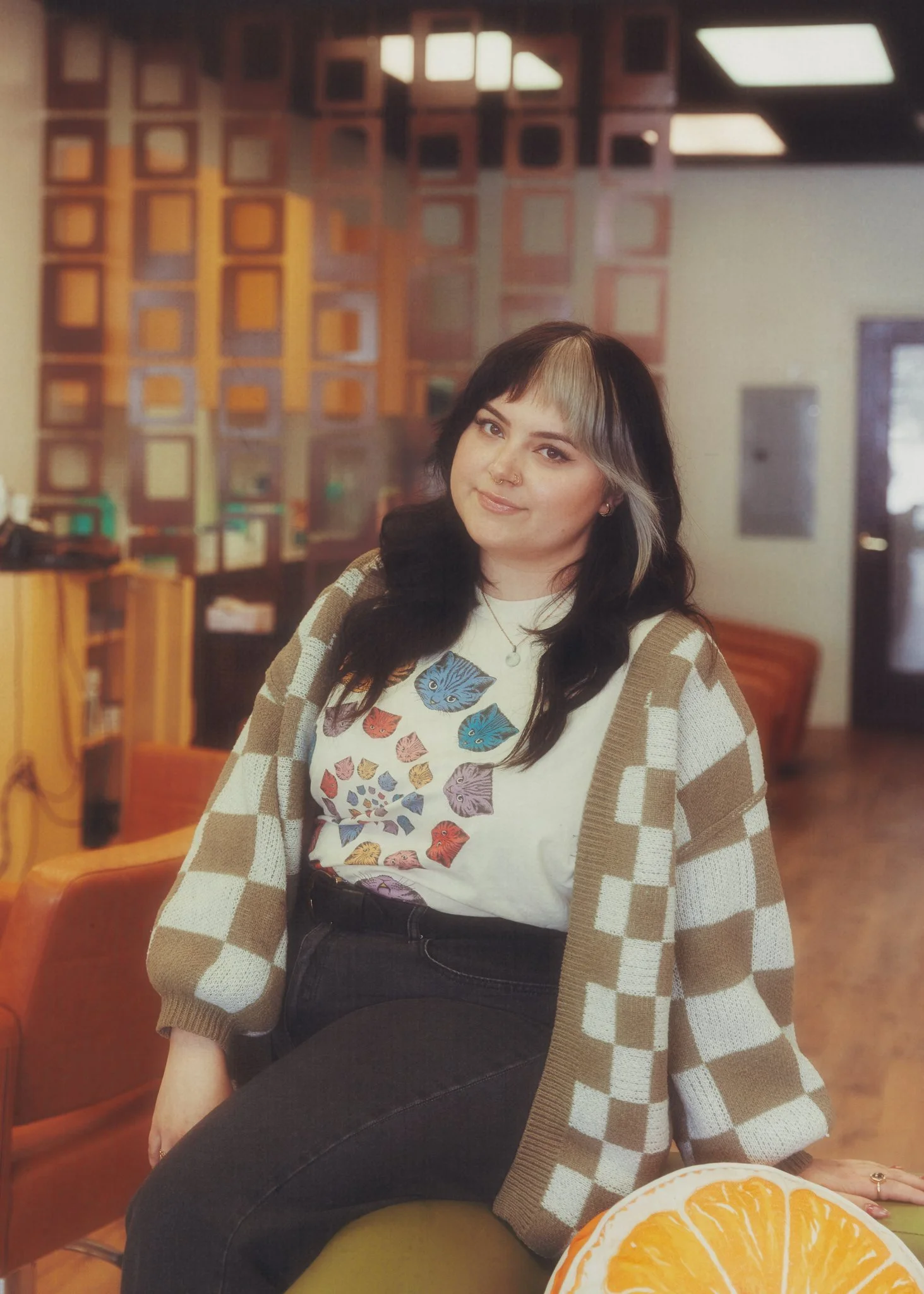 Kaila poses in the salon with the washing station behind her. She has medium length dark hair with a blonde section in her bangs. She's wearing a graphic t-shirt with a brown and white checkered sweater and black jeans.