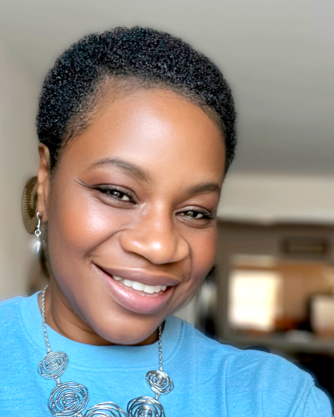 A smiling woman with short curly hair, wearing a blue shirt, a silver floral necklace, and a pearl earring, indoors.