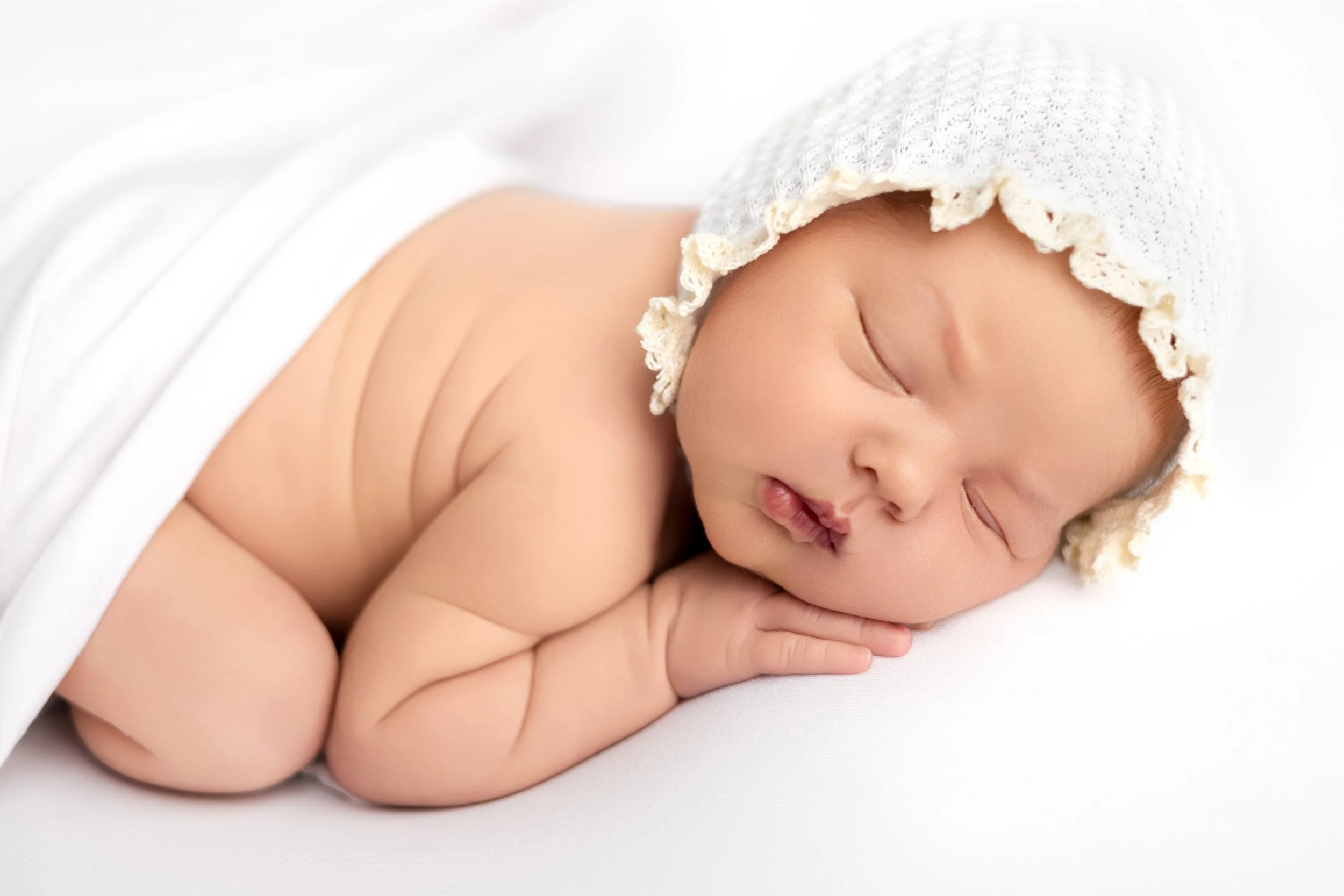 A sleeping newborn baby with a knitted hat, lying on their side on a white surface with a white blanket partially covering them.