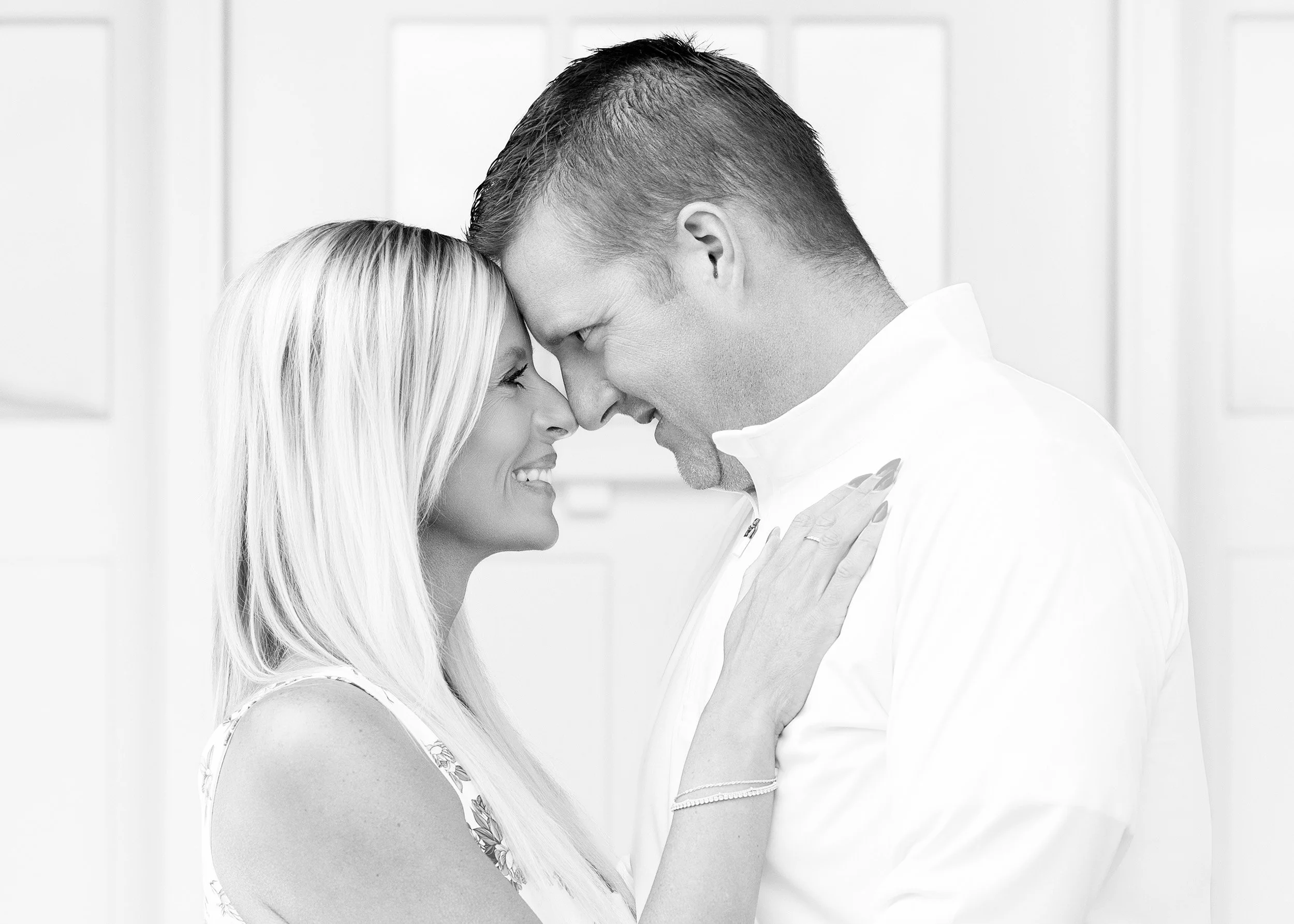 A black and white photograph of a couple with their foreheads touching, eyes closed, smiling, in an intimate moment, indoors with a blurred background.