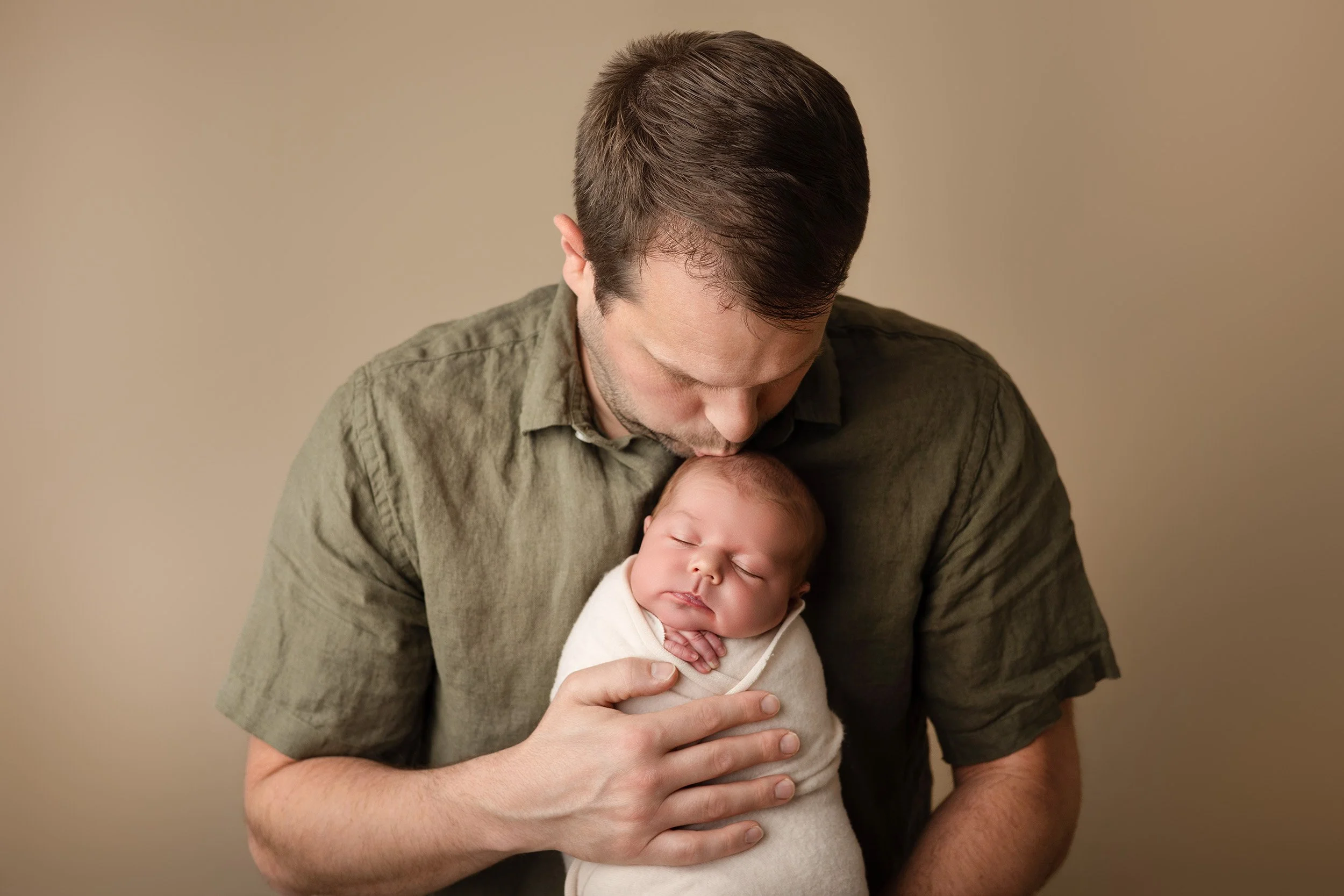 A man with dark hair and a beard holding a sleeping baby with blonde hair, against a neutral beige background.