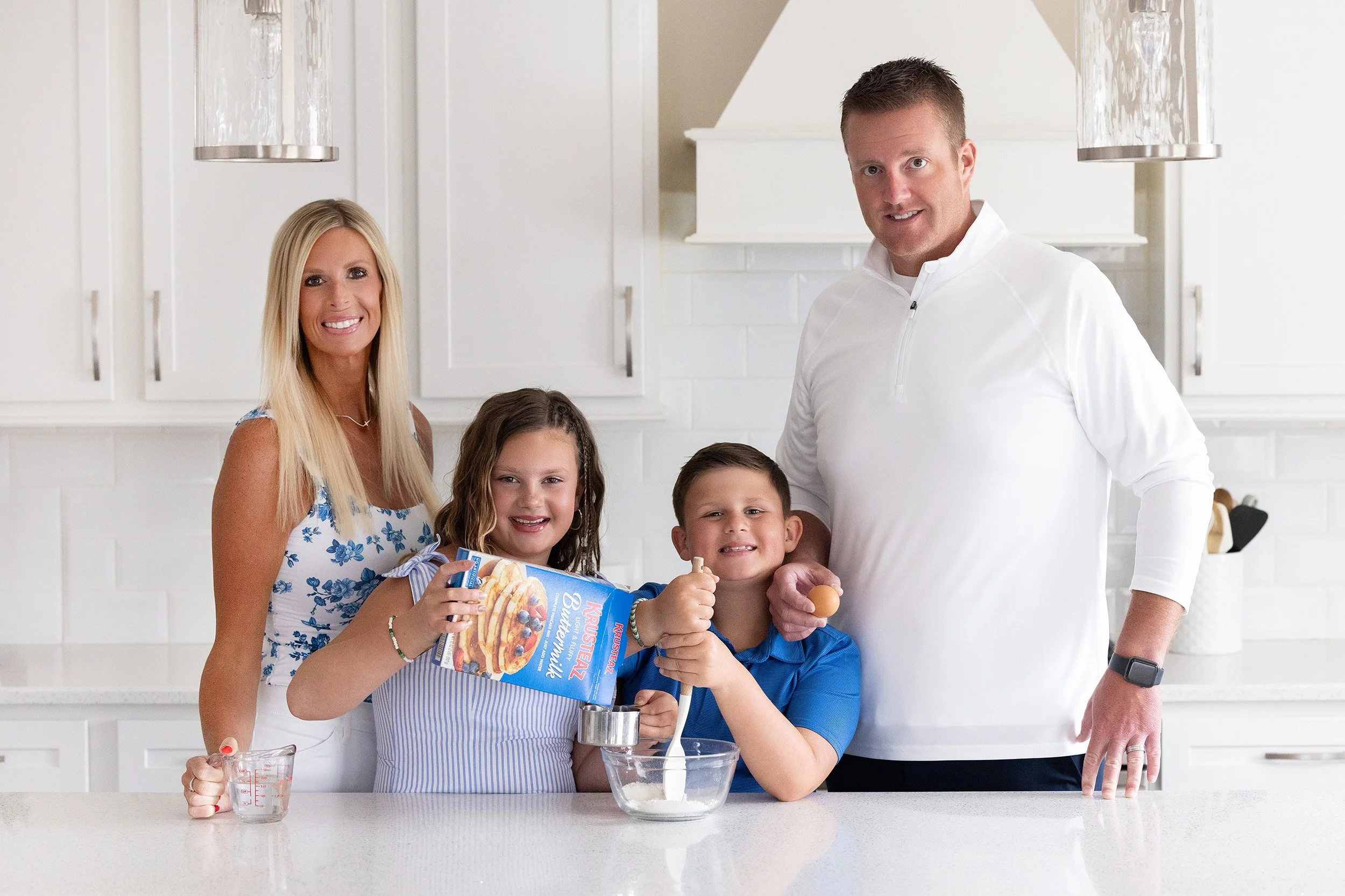 Family of four in a kitchen baking together, smiling at the camera, with ingredients and baking tools on the counter.