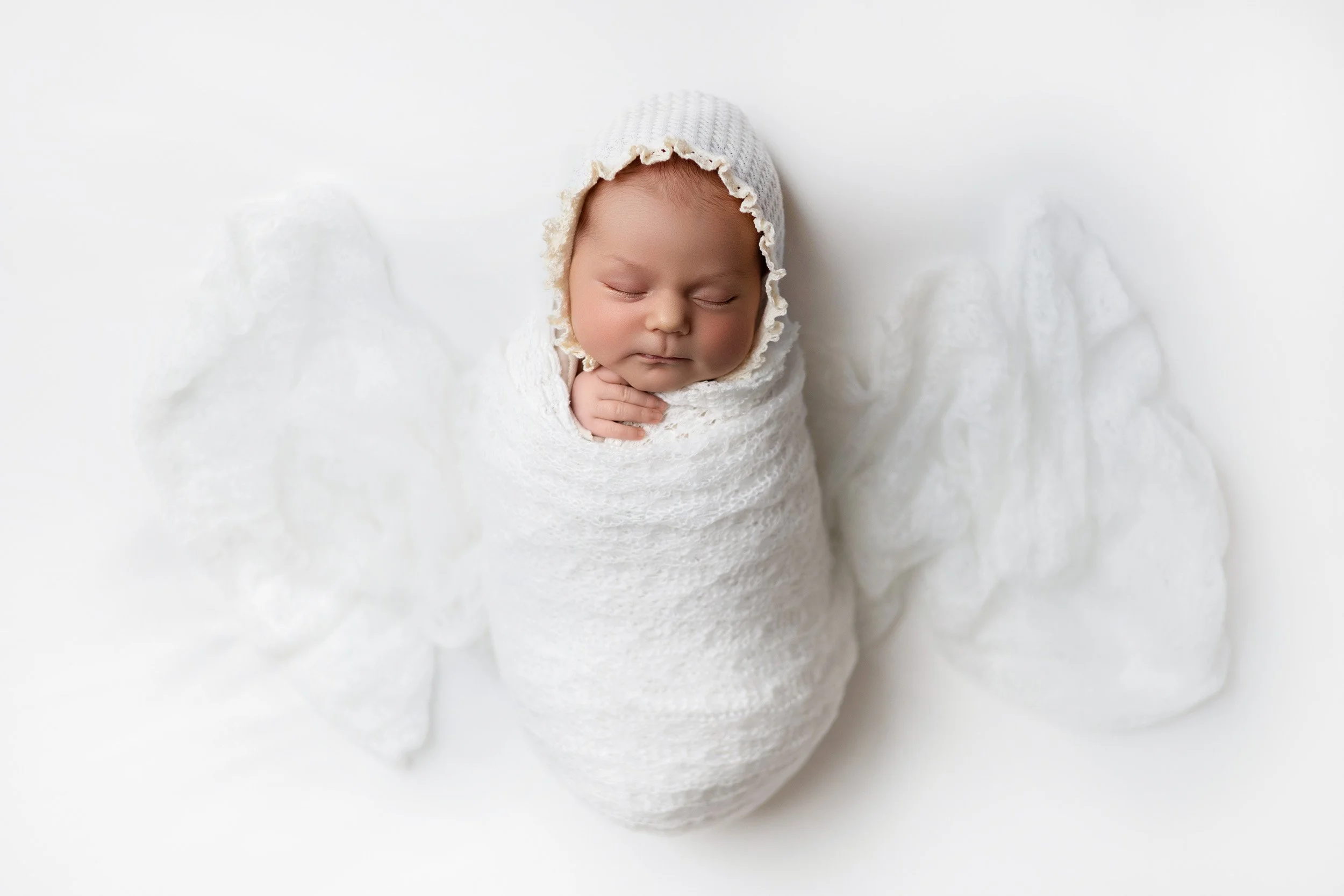 Close-up of a sleeping infant wrapped in white cloth, wearing a white knitted bonnet, against a white background.