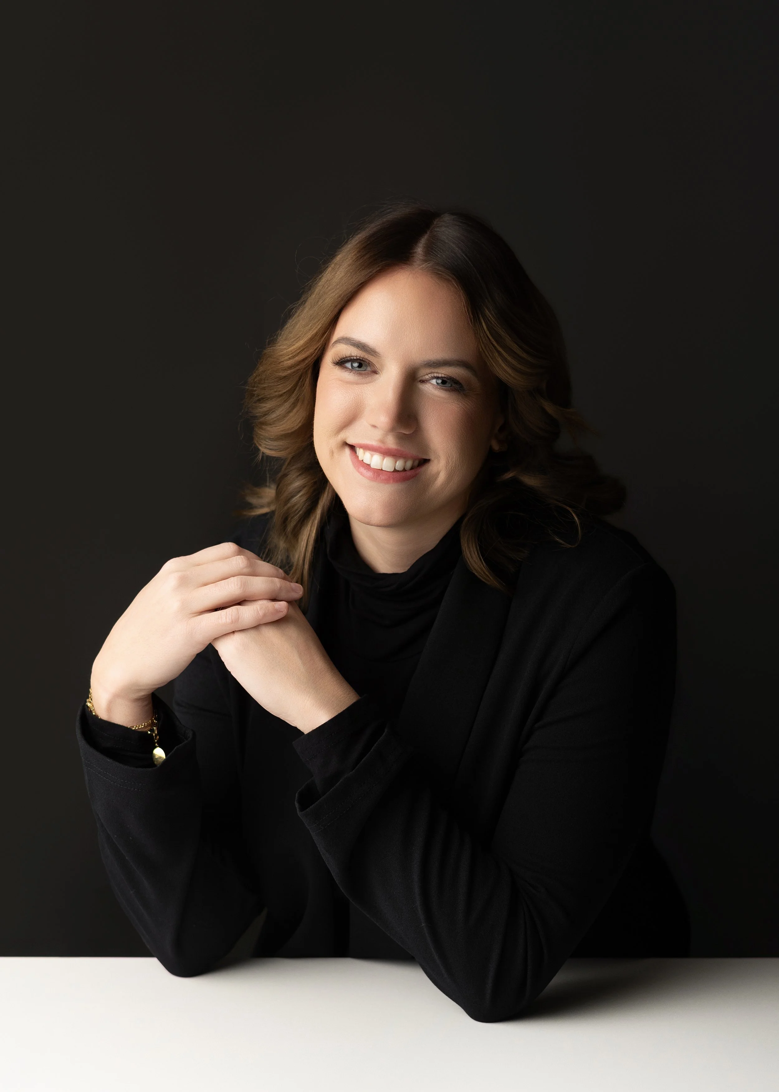 A woman with wavy brown hair smiling and sitting at a white table against a dark background, wearing a black shirt.