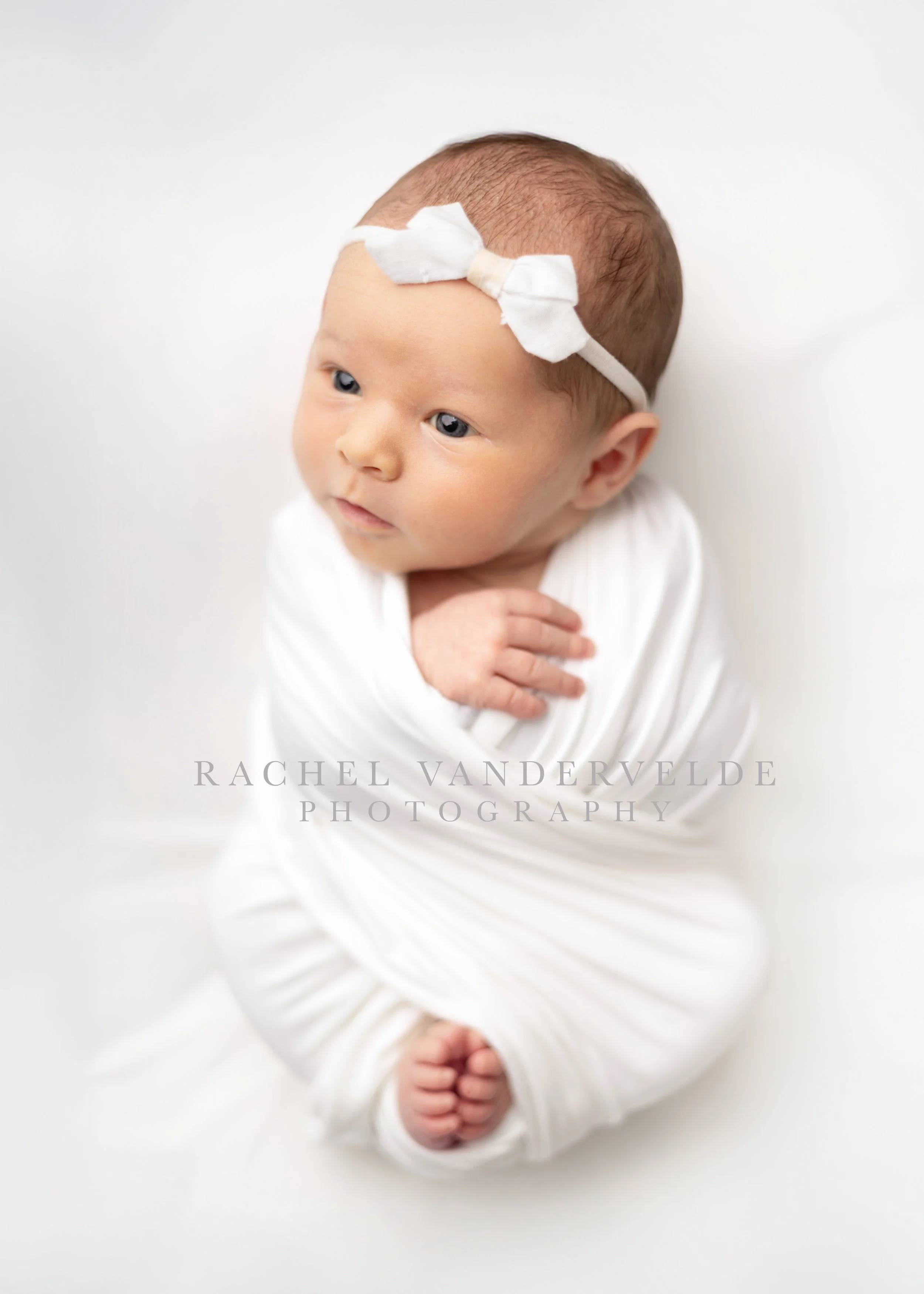 A baby with a white bow on the forehead, lying on a white blanket, with hands clasped. The newborn has light skin, blue eyes, and light brown hair.