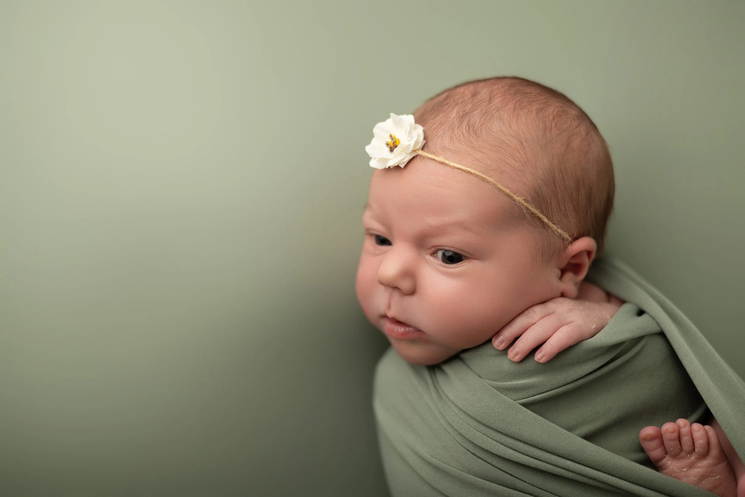 Close-up of a baby with a flower headband, resting on their arm, wearing a green outfit, against a light green background.