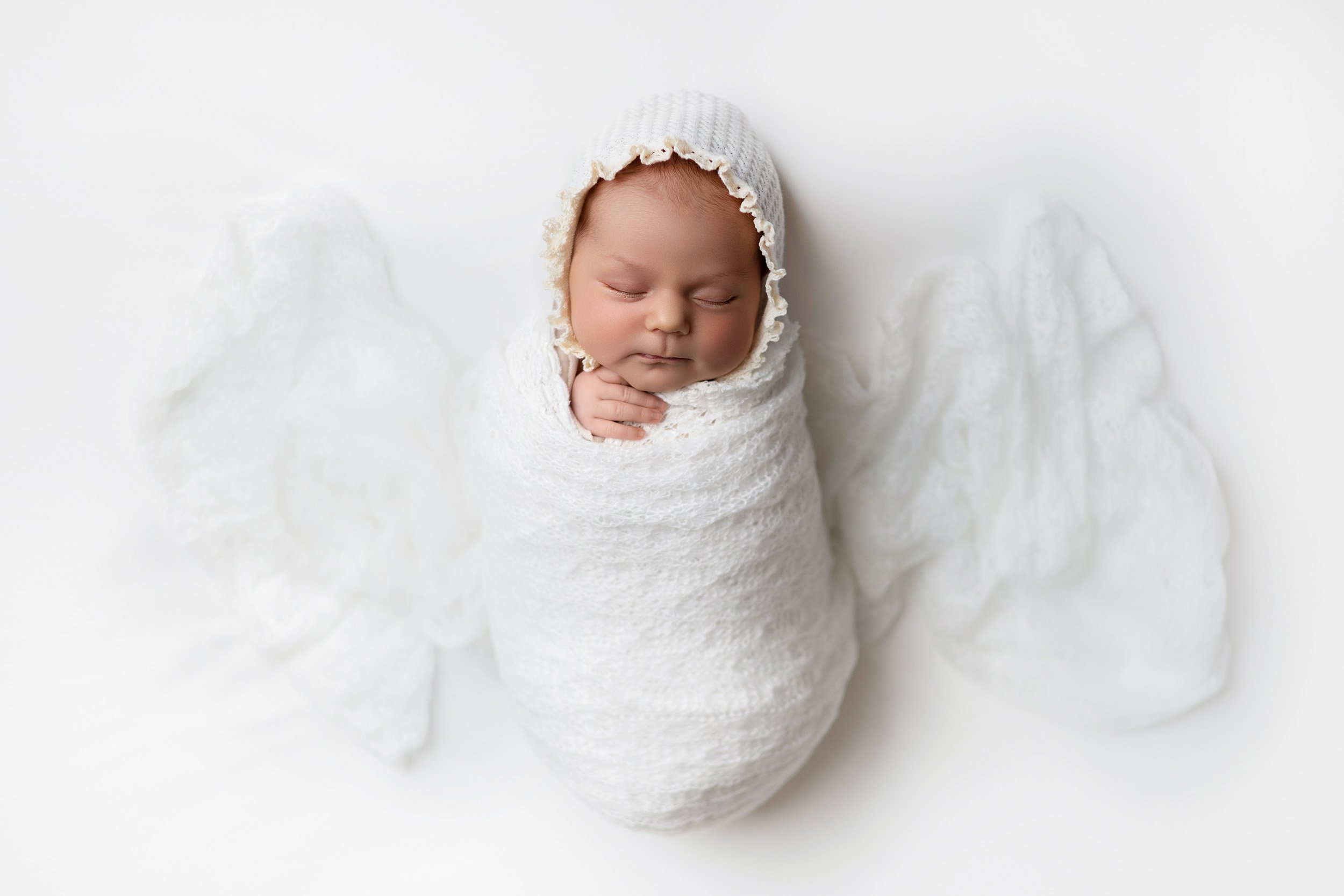 Close-up photo of a newborn baby in a knitted bonnet, wrapped in a white blanket, peacefully sleeping on a white surface.
