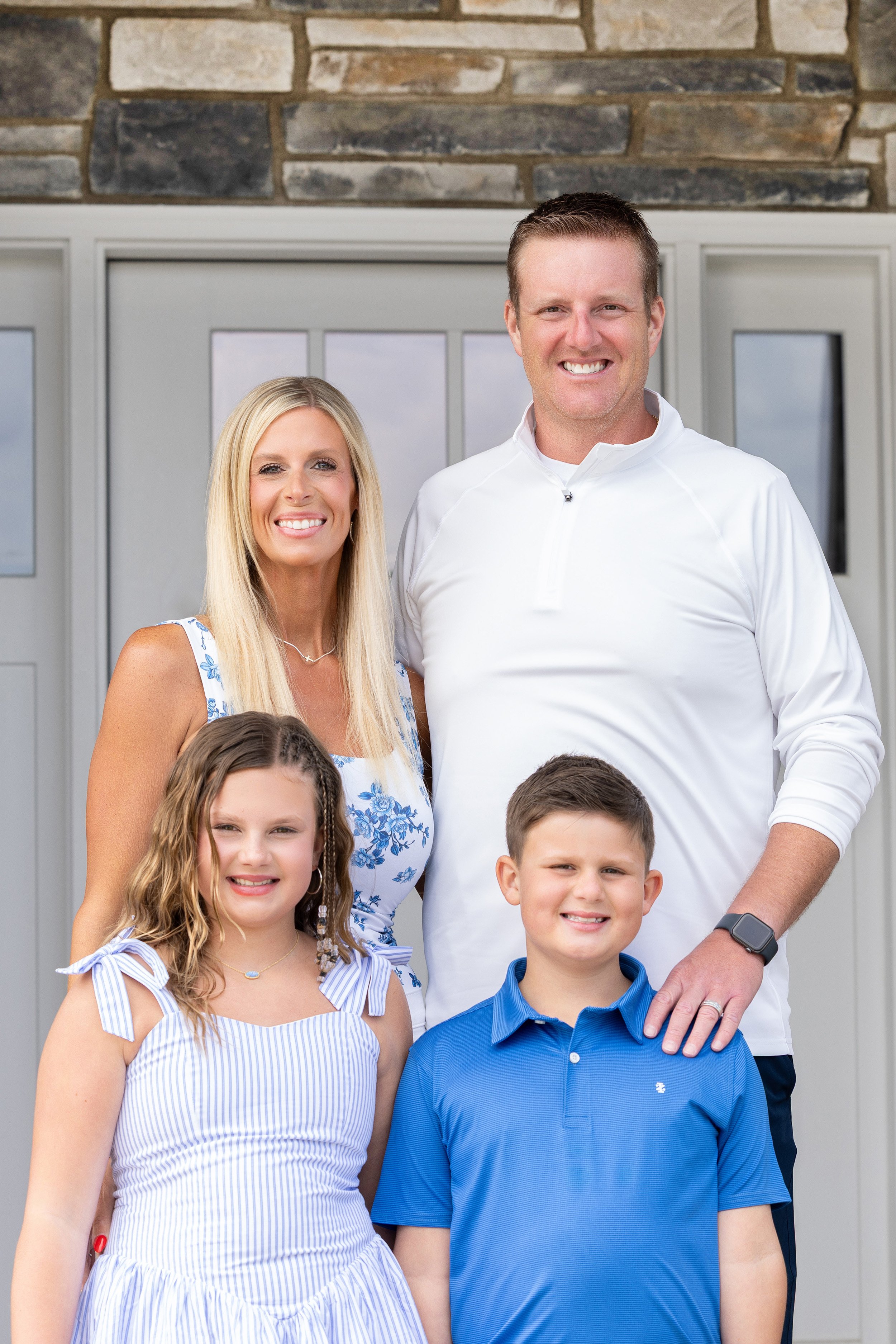 Group portrait of a smiling family standing in front of a house with a brick and siding exterior. The group includes two adults and four children, all dressed in casual summer clothing.