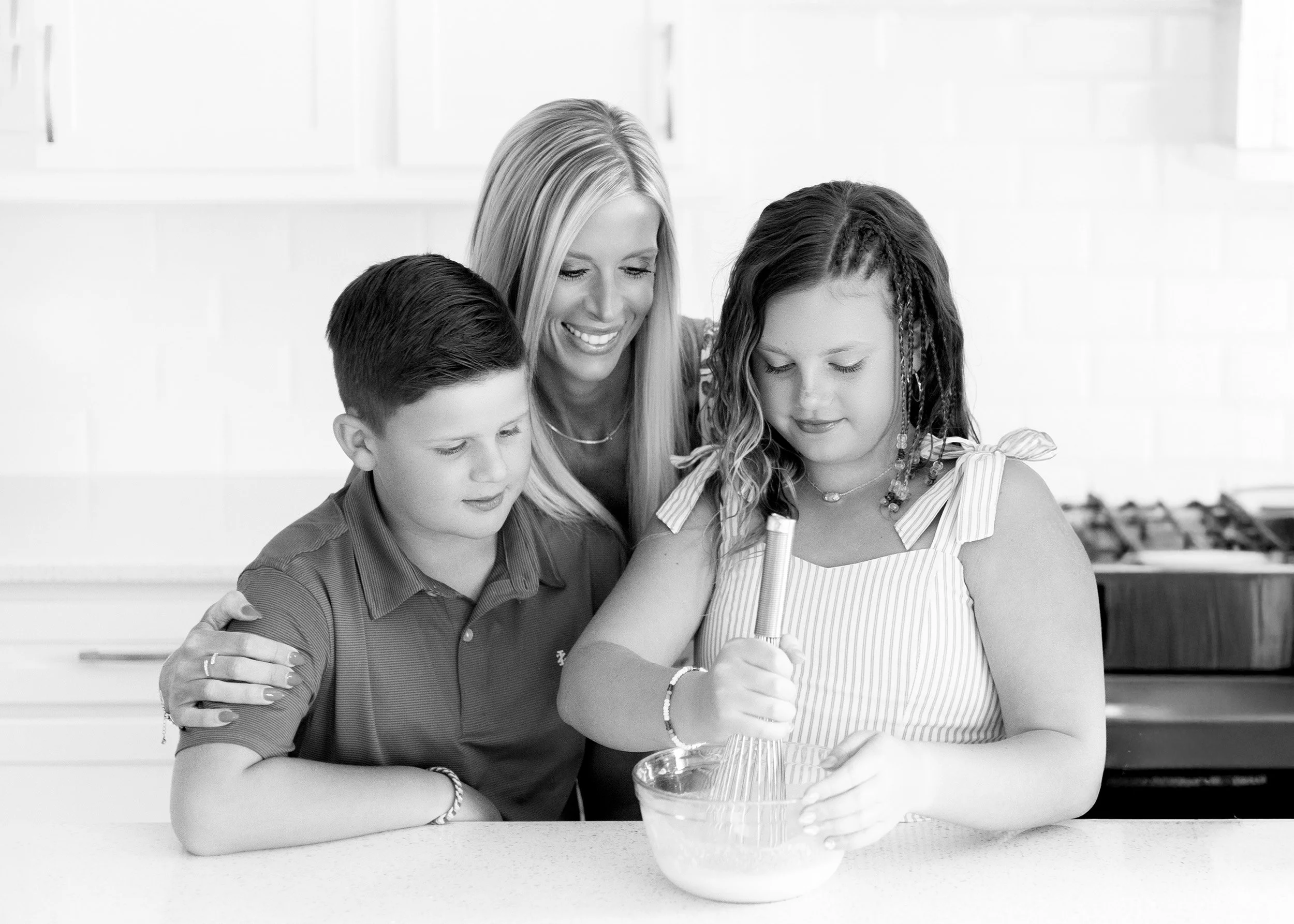 A woman and two children sitting at a kitchen counter, with the woman helping one of the children make a cake.