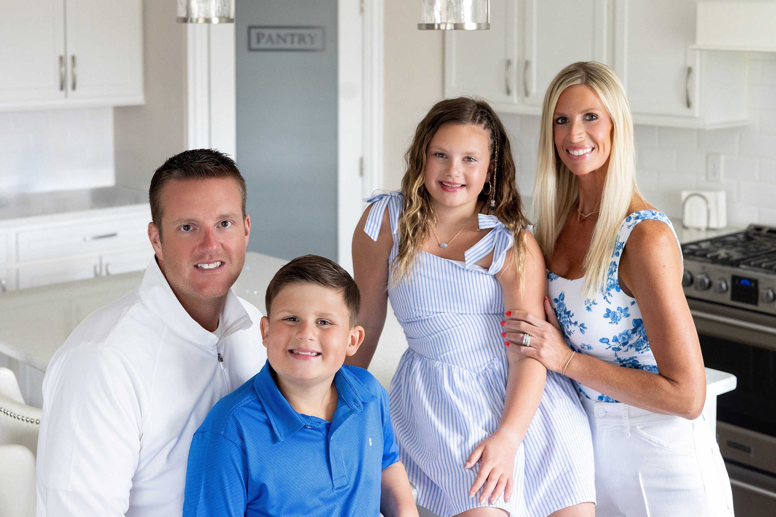 A family of four posing in a modern white kitchen. The father and mother are standing behind two children, a boy and a girl. All family members are smiling and dressed in blue and white clothing.