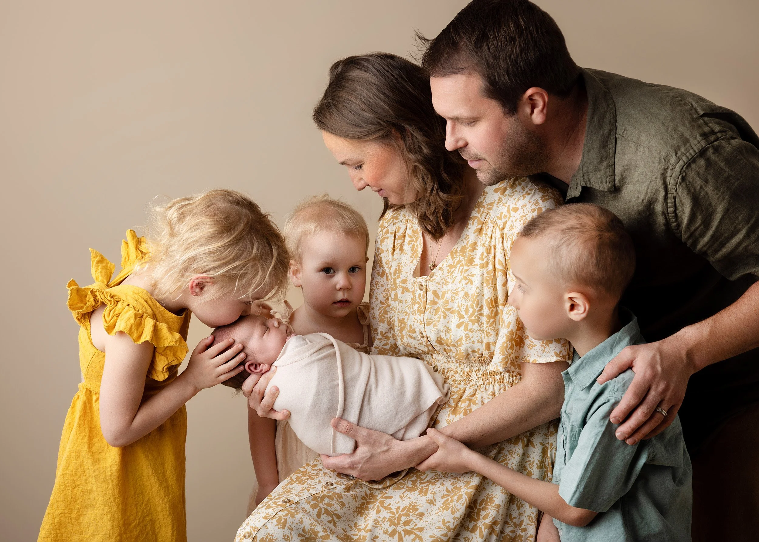 A family gathers around a newborn baby, with parents and young children affectionately holding and looking at the baby, indoors against a plain beige wall.