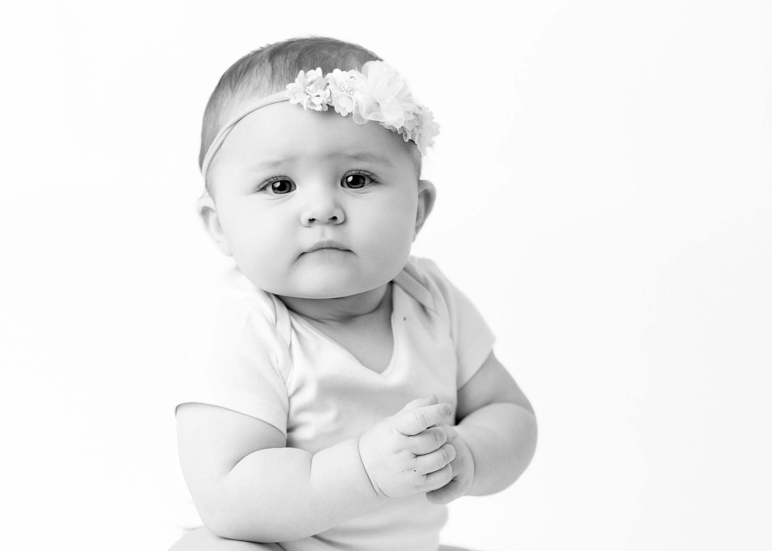 Black and white photo of a baby girl with big eyes and chubby cheeks wearing a floral headband and a short-sleeved shirt, with hands clasped together.