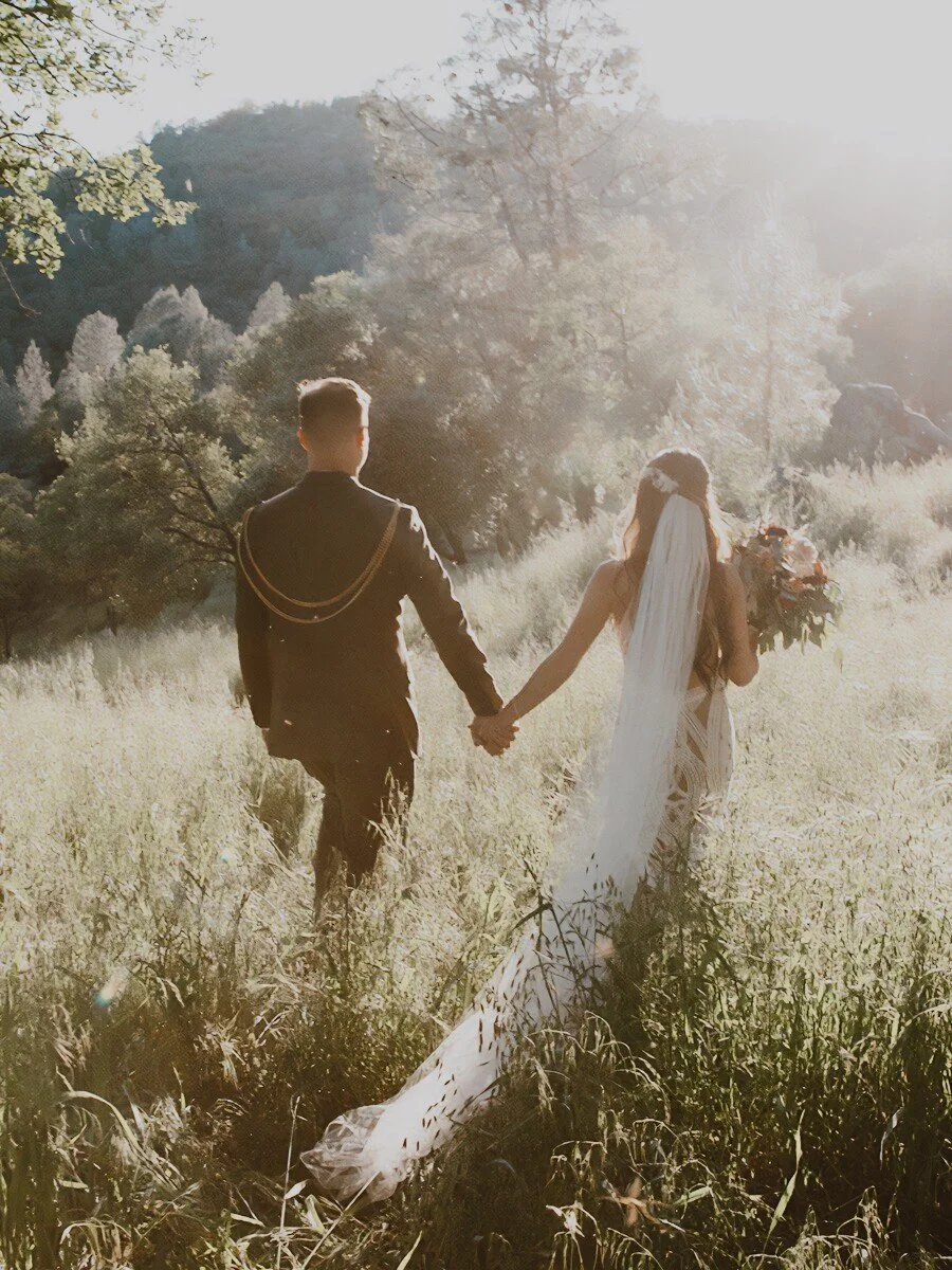 Wedding bride and groom walking hand in hand through lush grassy landscape with sun setting in the west