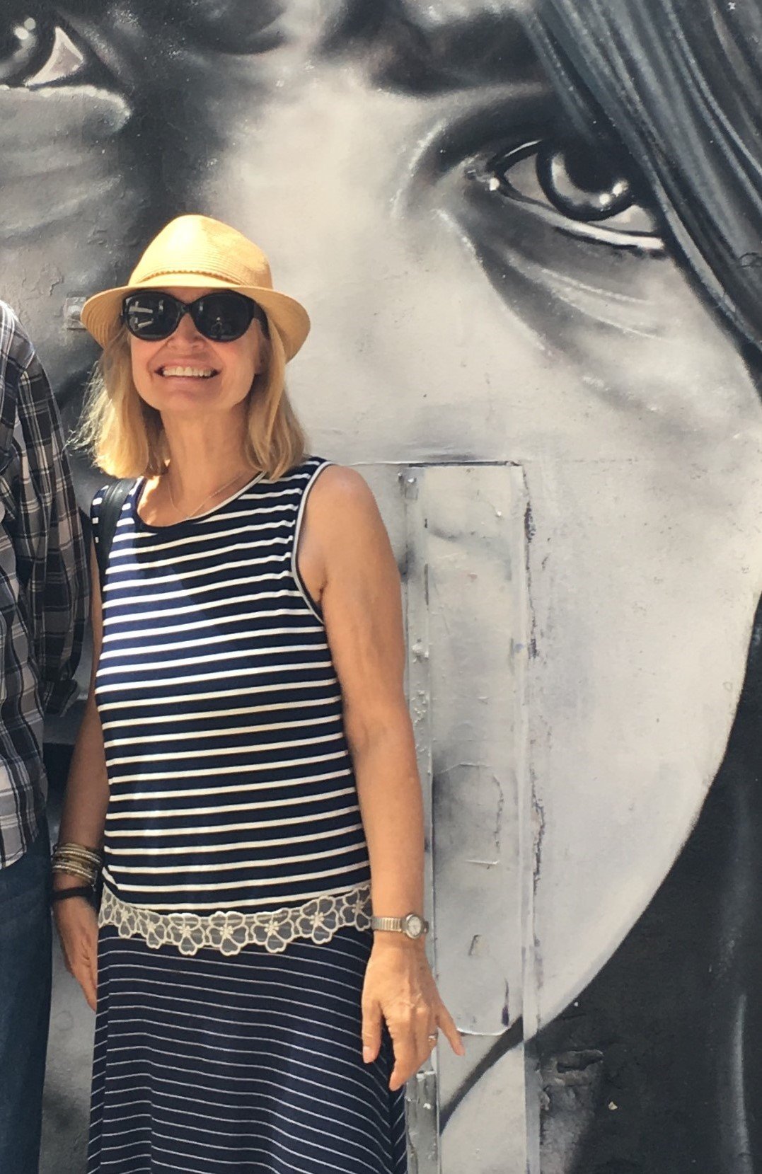 Smiling woman wearing sunglasses, a straw hat, and a navy and white striped dress standing in front of a black and white mural of a person's face.