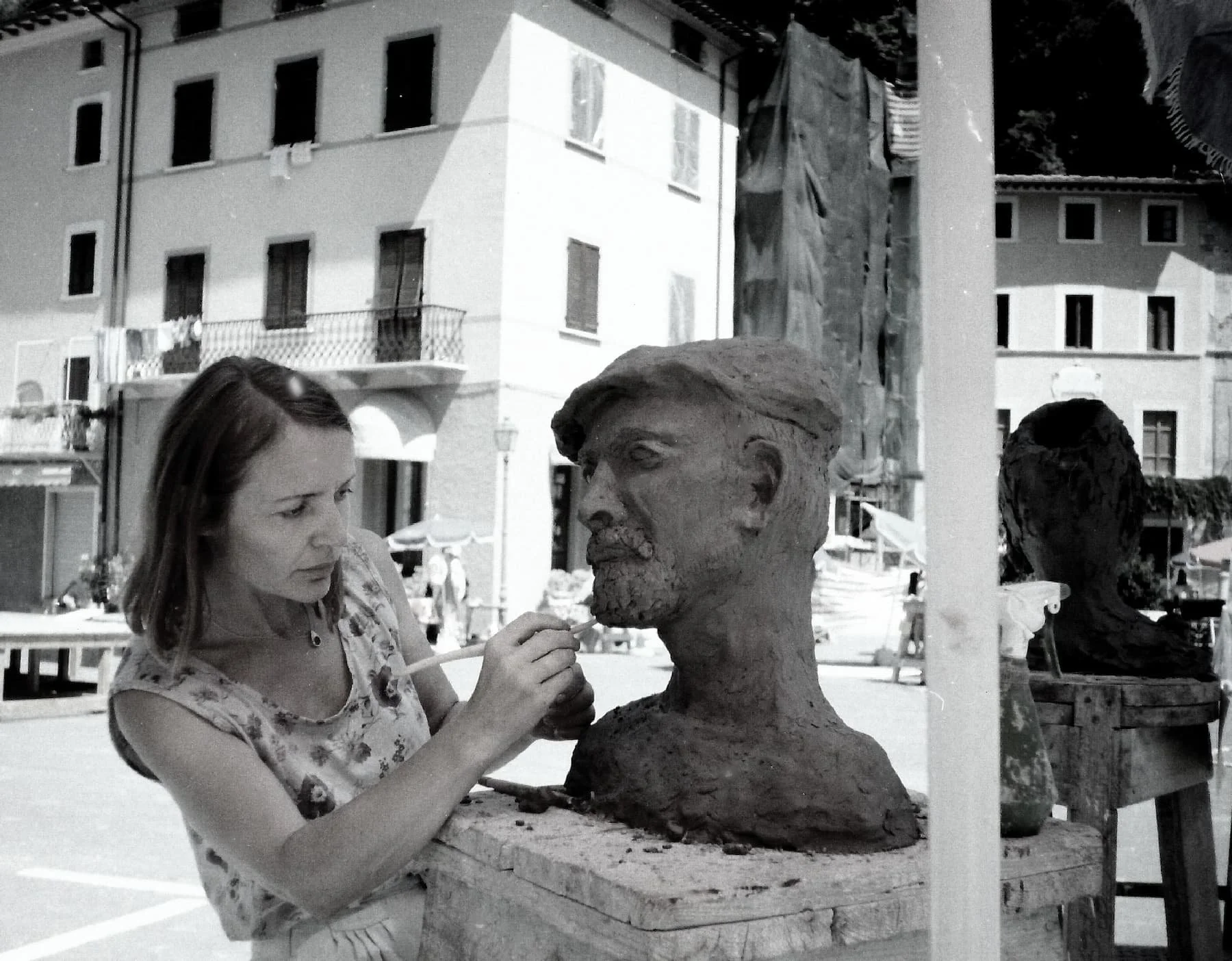 A woman working on a clay sculpture of a man's bust outdoors in a European town square. Buildings with balconies and laundry hanging can be seen in the background.