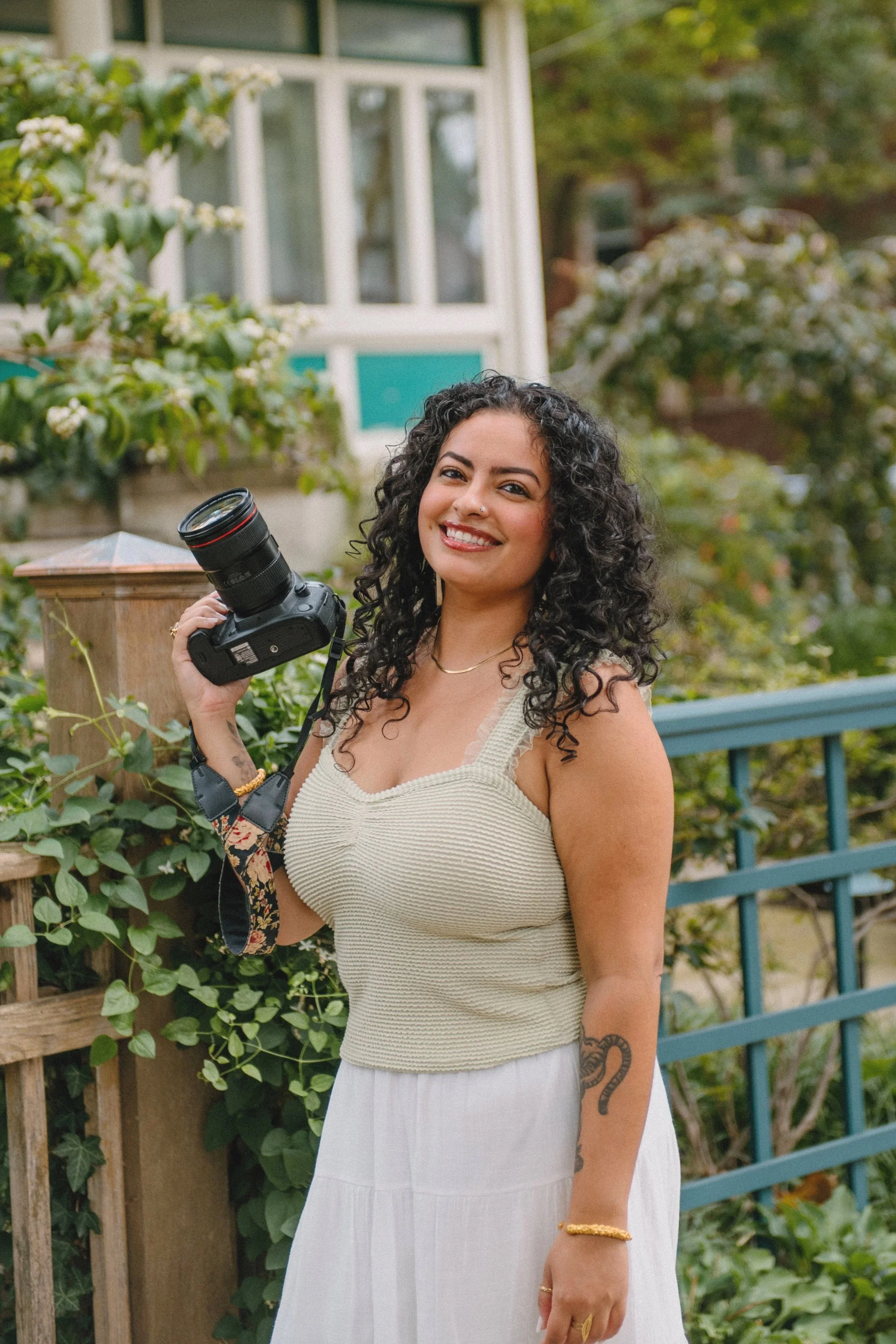 A woman standing by a house, holding a camera, wearing a light green top and long white skirt