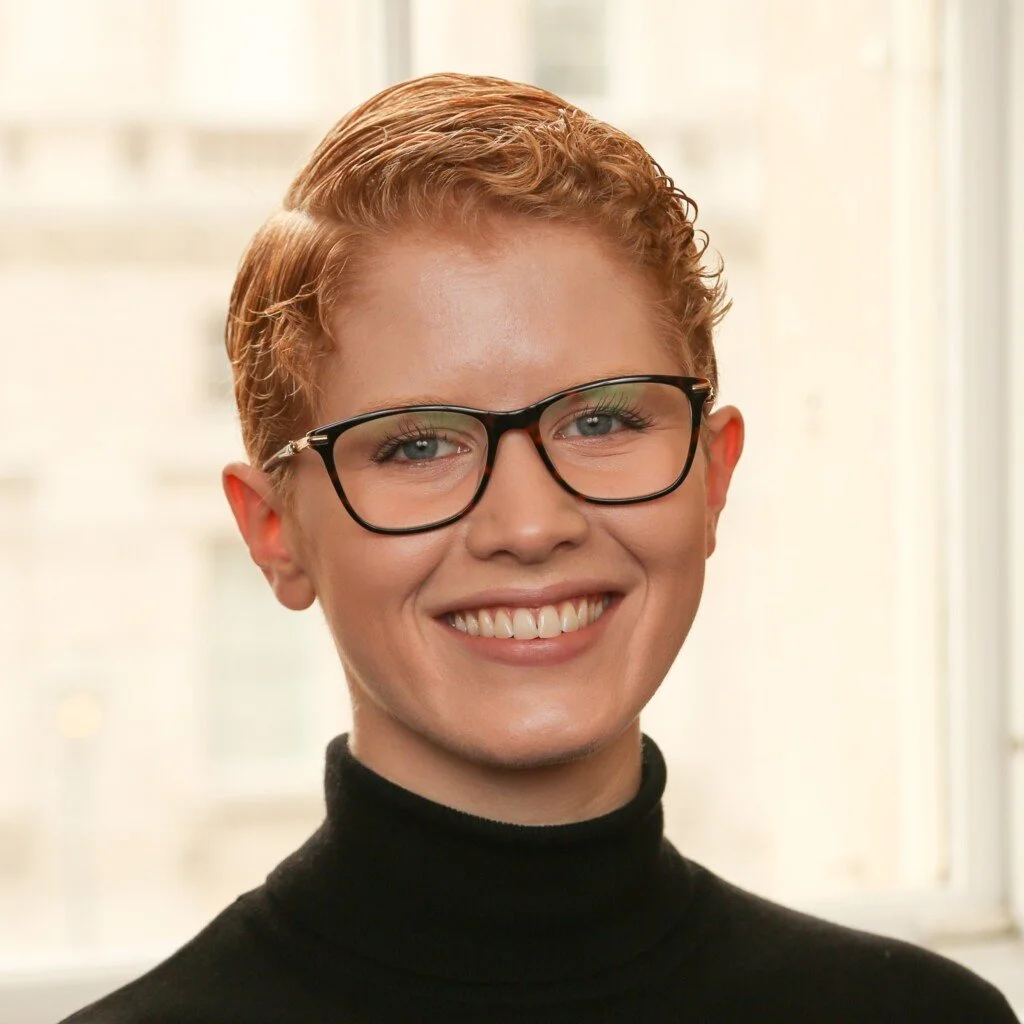 A woman with short curly red hair wearing black glasses and a black turtleneck, smiling in front of a bright window.