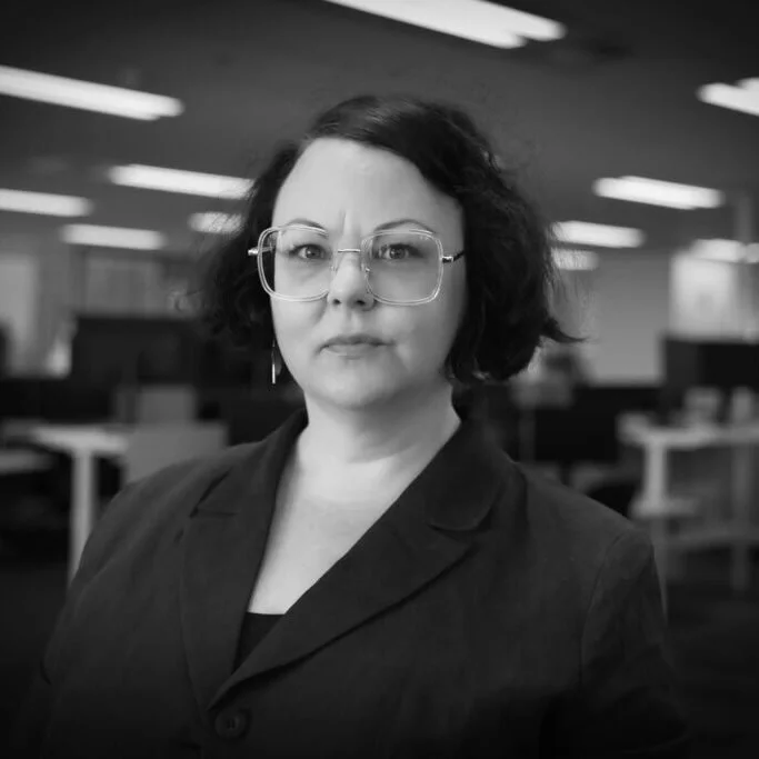 A woman with short wavy dark hair, wearing large clear eyeglasses, a nose piercing, and a dark blazer, standing in an office with blurred computer desks and overhead lighting.