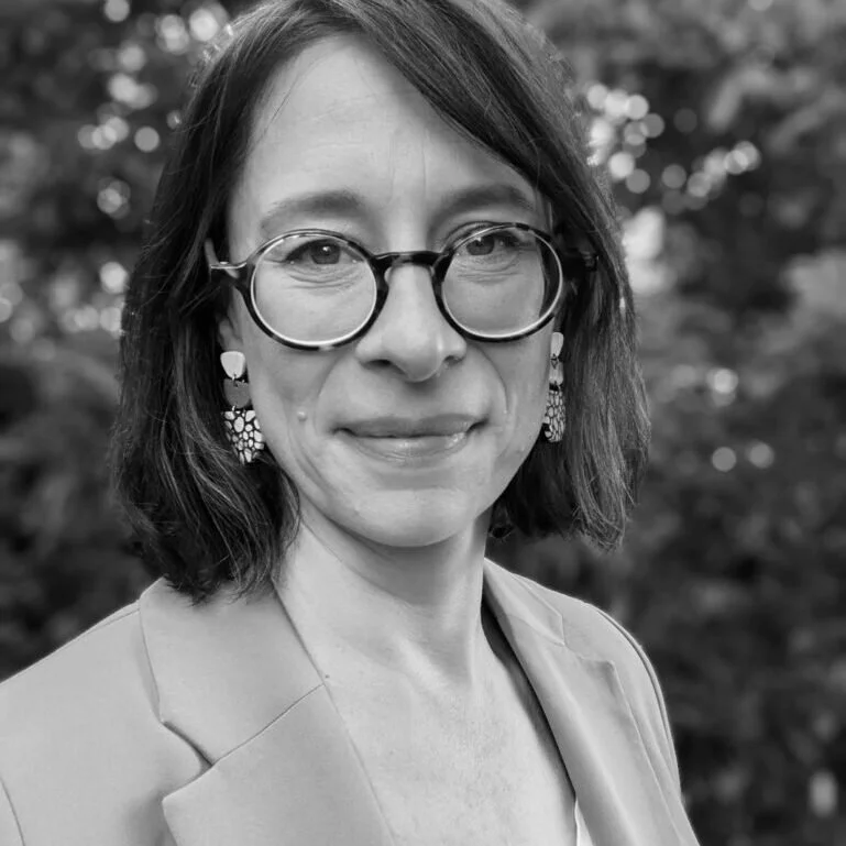 Black and white portrait of a woman wearing glasses, earrings, and a blazer, standing outdoors with blurred trees in the background.