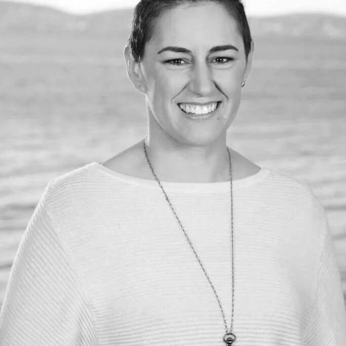 A woman with short dark hair smiling, wearing a light-colored top and a long necklace, standing outdoors near a body of water.