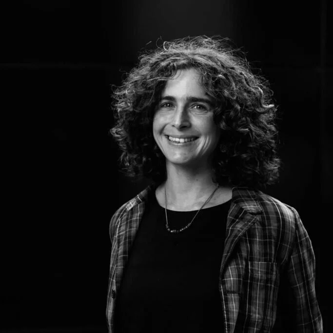 A woman with curly hair smiling, wearing a plaid shirt and necklace, against a dark background.