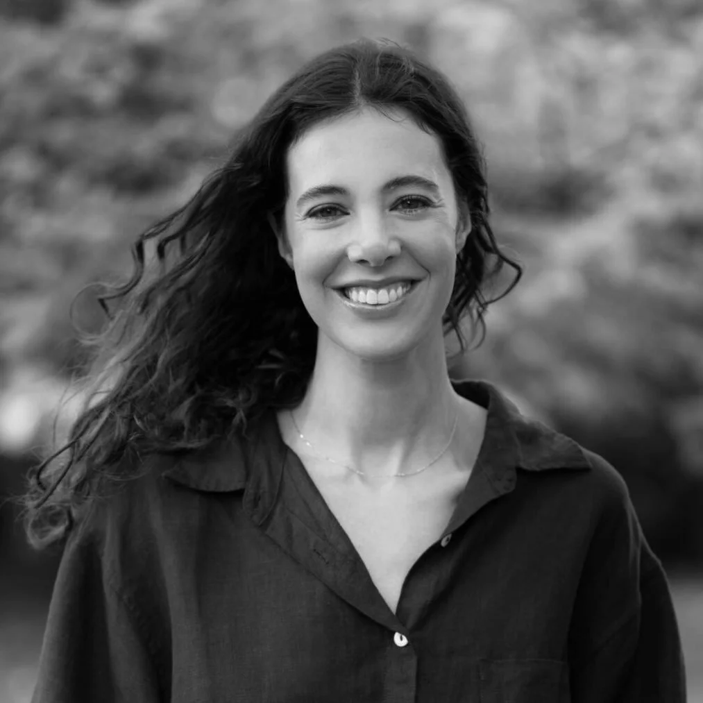 Black and white photo of a smiling woman with long, wavy hair, wearing a dark shirt outdoors.