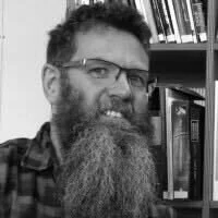 A man with glasses and a beard, smiling, in front of a bookshelf