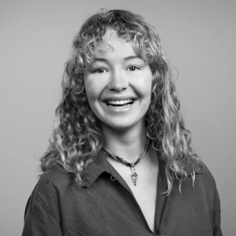 Black and white portrait of a smiling woman with curly hair, wearing a button-up shirt and a necklace, against a plain background.