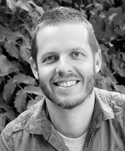 A smiling man with short hair and a beard outdoors, standing in front of leafy plants.