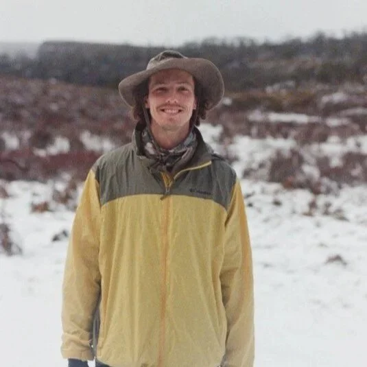 A man standing outdoors in a snowy landscape, wearing a wide-brimmed hat, a yellow and gray jacket, and smiling at the camera.