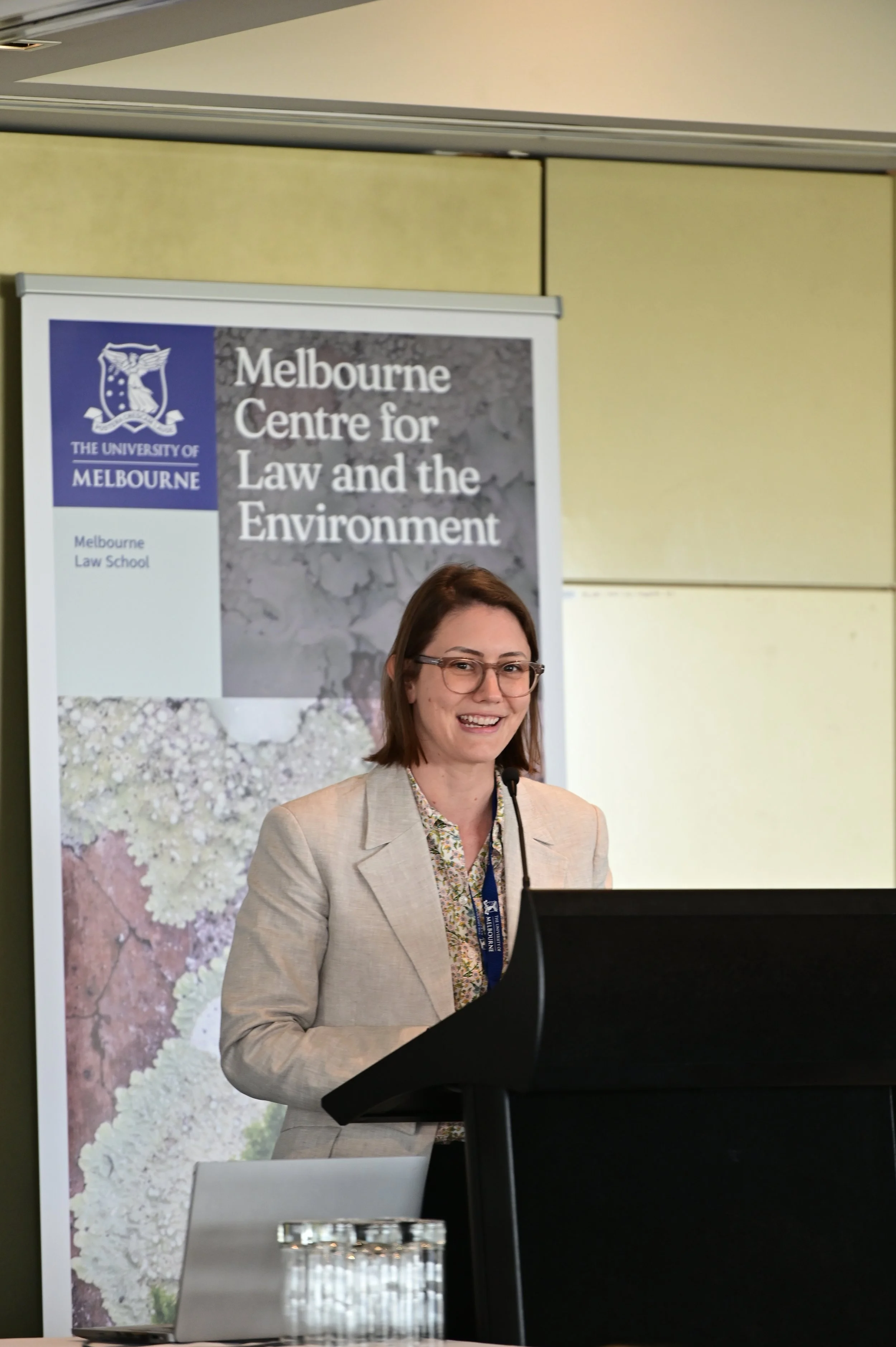 A woman wearing glasses and a beige blazer is standing at a podium, speaking at an event for the University of Melbourne Center for Law and the Environment. There is a large banner behind her with the university's logo and the event title.