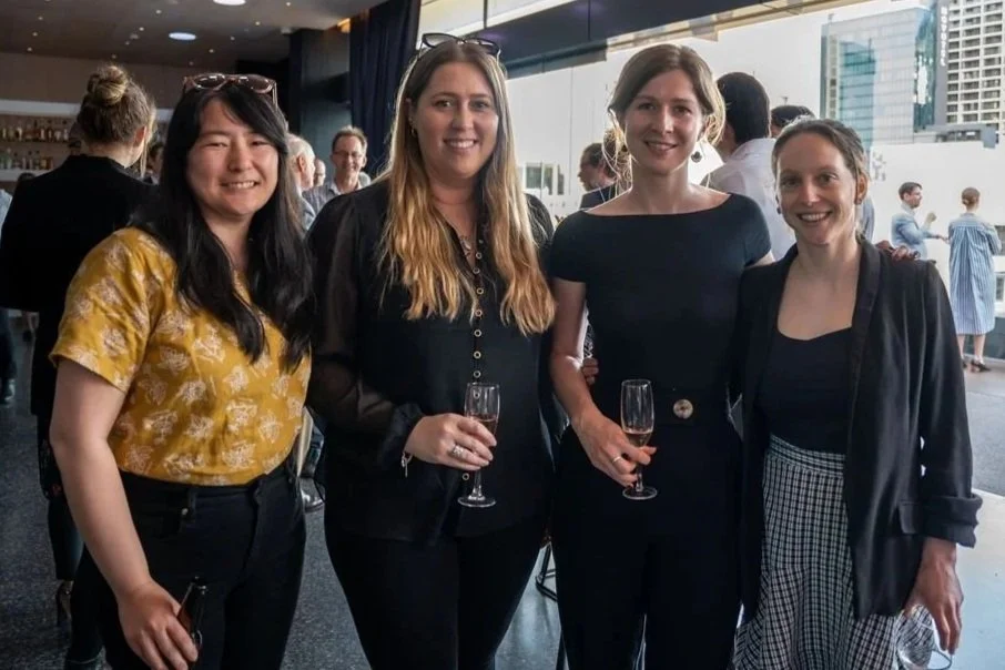 Four women standing together at a social event, holding champagne glasses, in a modern indoor space with large windows and city buildings outside.