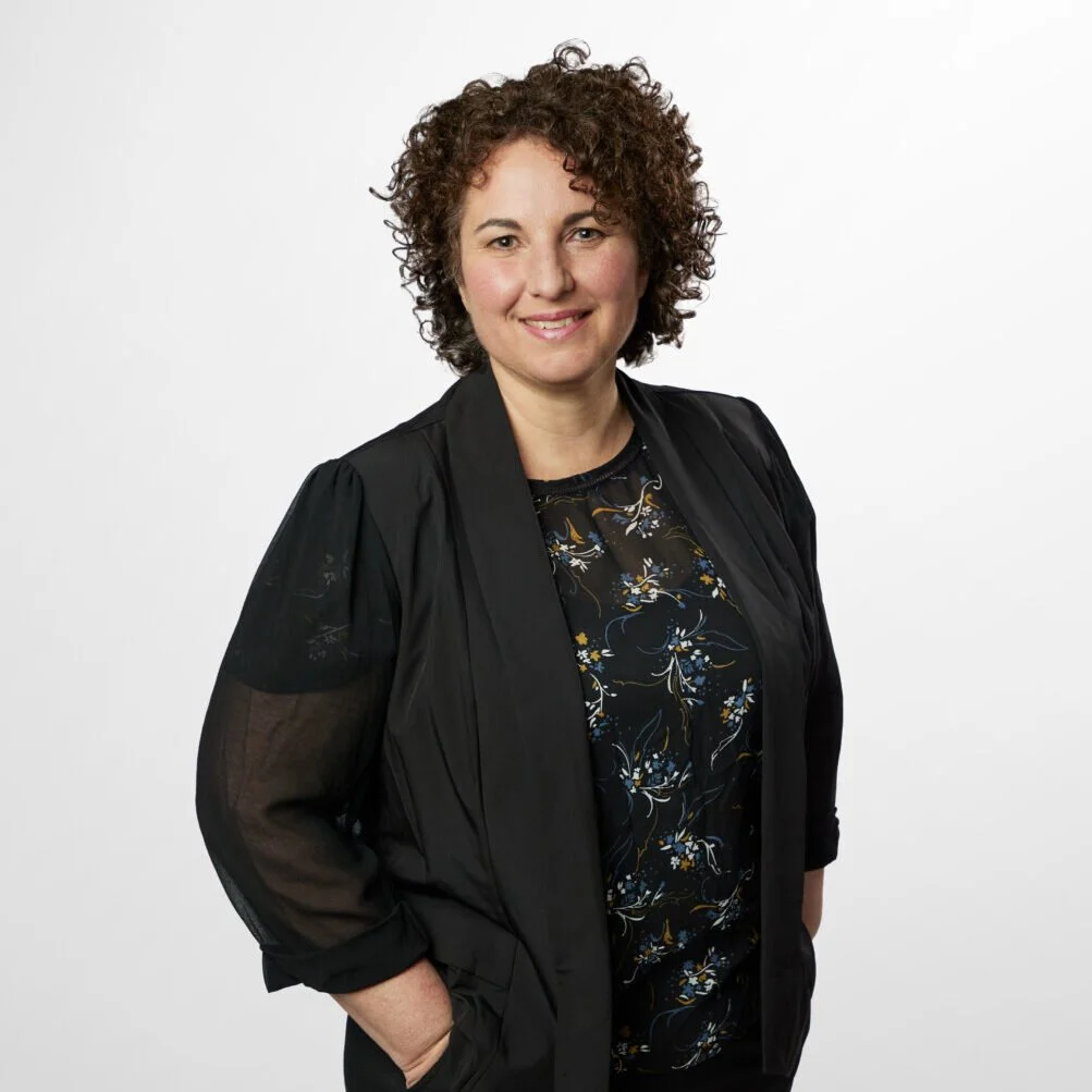 A woman with curly brown hair smiling, wearing a black jacket over a patterned shirt, standing against a plain white background.