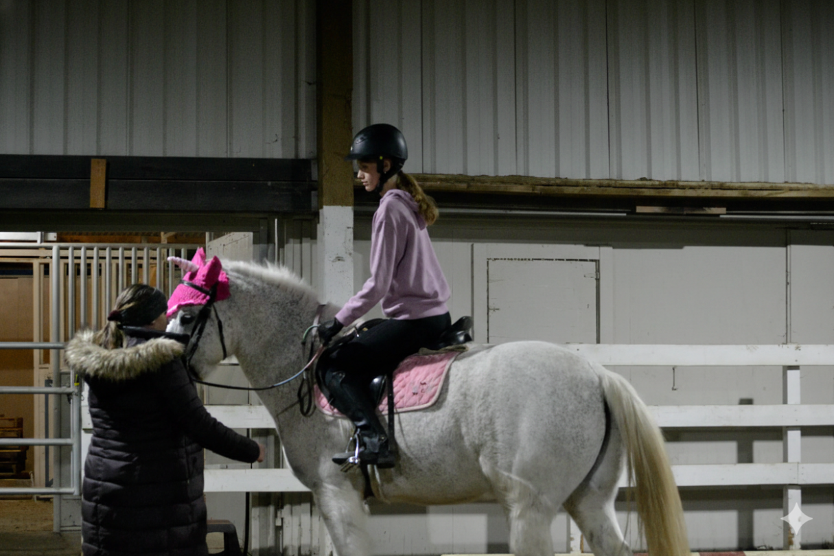 A girl wearing a pink hoodie and black riding helmet sitting on a white horse inside an indoor riding arena, with Theresa in a black coat adjusting the horse's bridle.