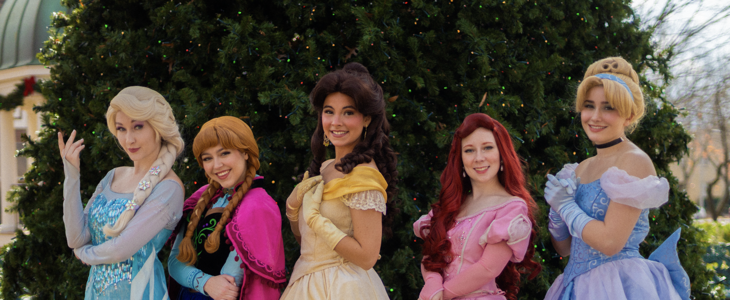 Five women dressed as Disney princesses standing in front of a decorated Christmas tree, smiling and posing outdoors.