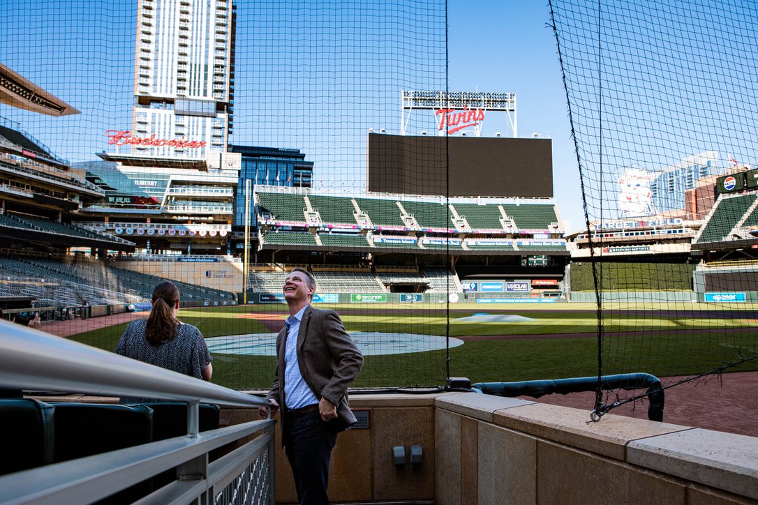 A man in a suit smiling and a woman with her back turned, standing in a baseball stadium near the field with empty seating in the background.