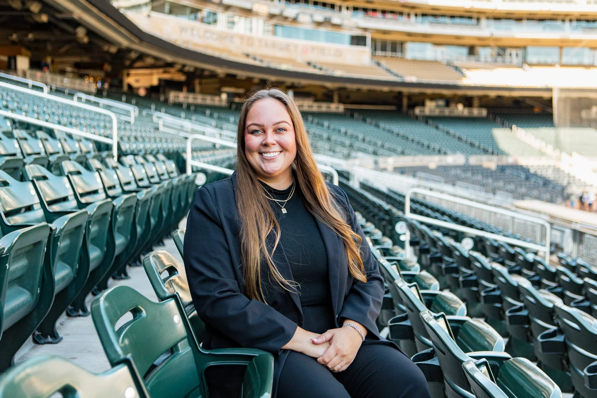 Young woman with long brown hair, wearing a black blazer and jewelry, sitting in an empty stadium with green seats, smiling at the camera.