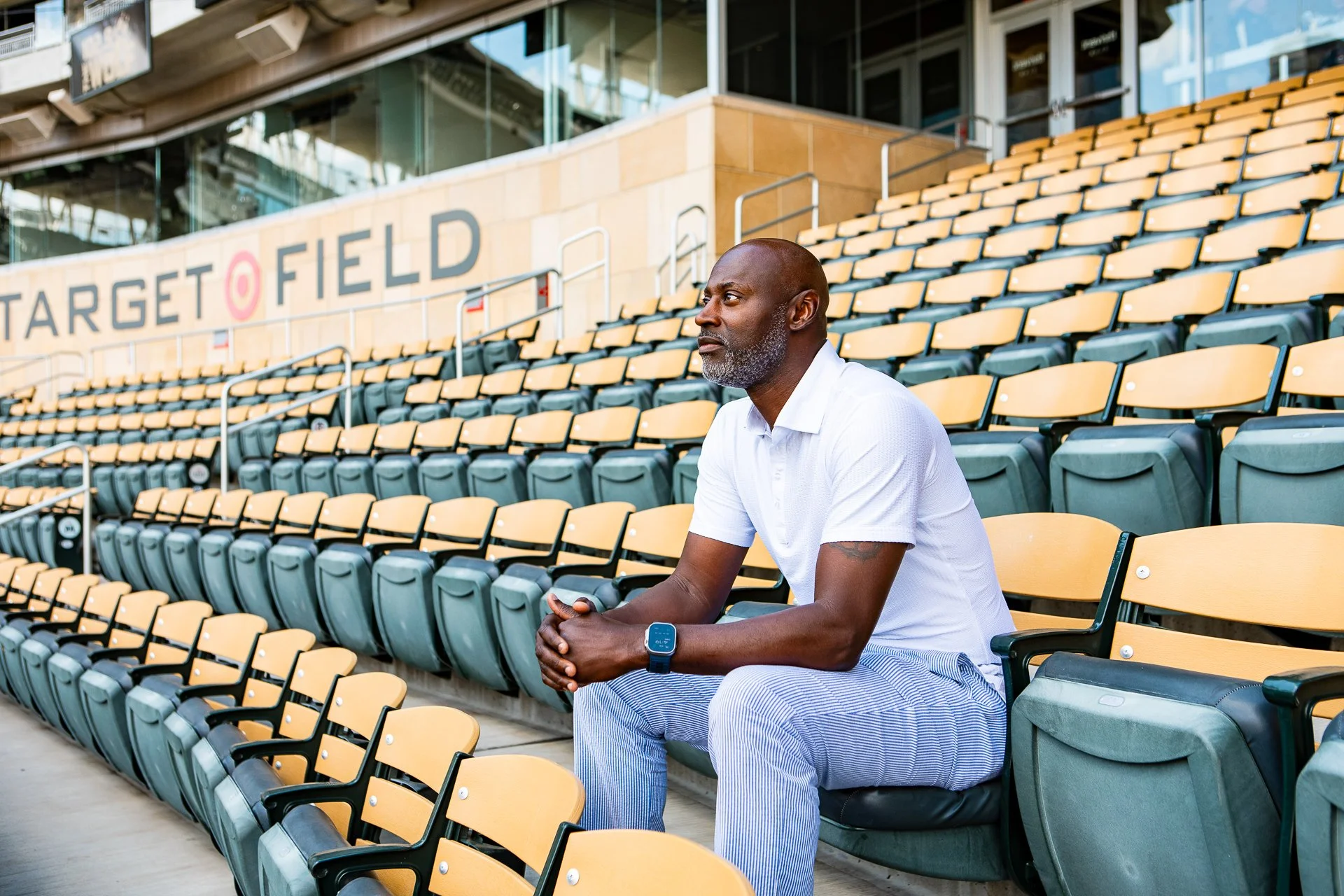 A man sitting alone on stadium seats at Target Field, wearing a white polo shirt, striped pants, and a smartwatch, holding a smartphone, with empty yellow and green seats behind him.