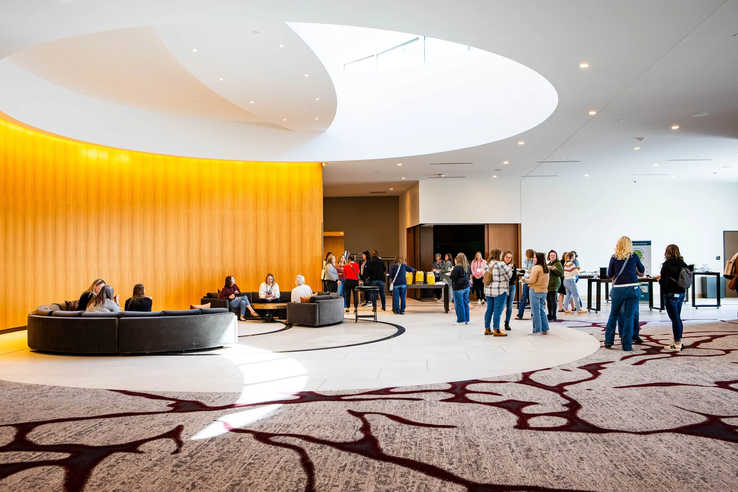 A spacious, modern indoor lobby with a high, curved ceiling and a large round skylight, filled with people waiting and socializing near a buffet or registration table, with a seating area featuring black curved couches and a patterned carpet.
