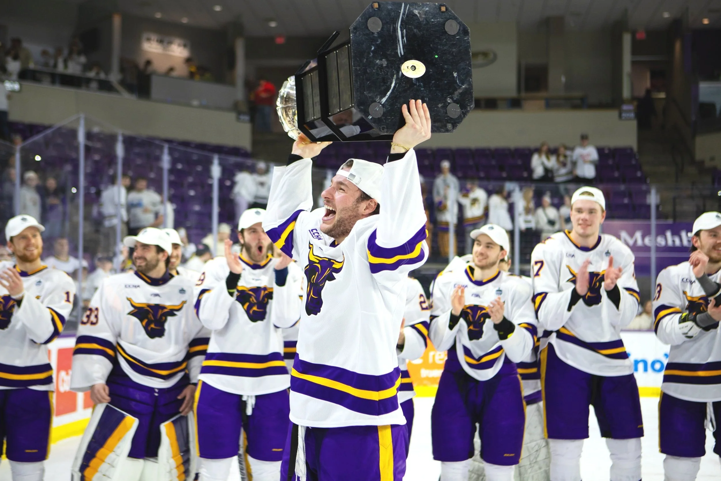 A hockey team celebrating on the ice after winning a championship, with one player holding a trophy above his head and others clapping and cheering.