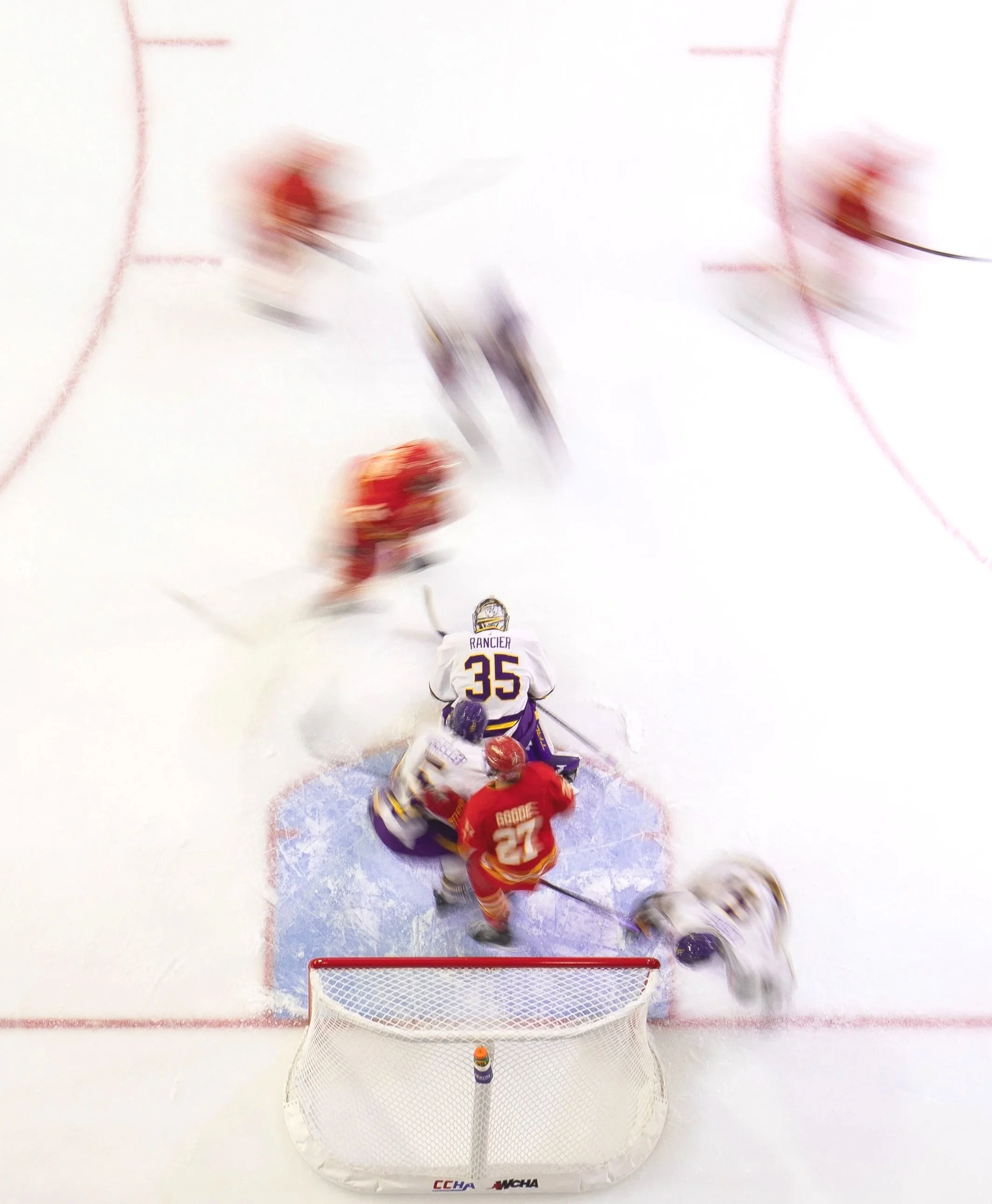 An Minnesota State University Mavericks ice hockey game with players around the crease, a goaltender in a white jersey with the number 35, and opposing players in red jerseys, one with the number 27, near the goal.