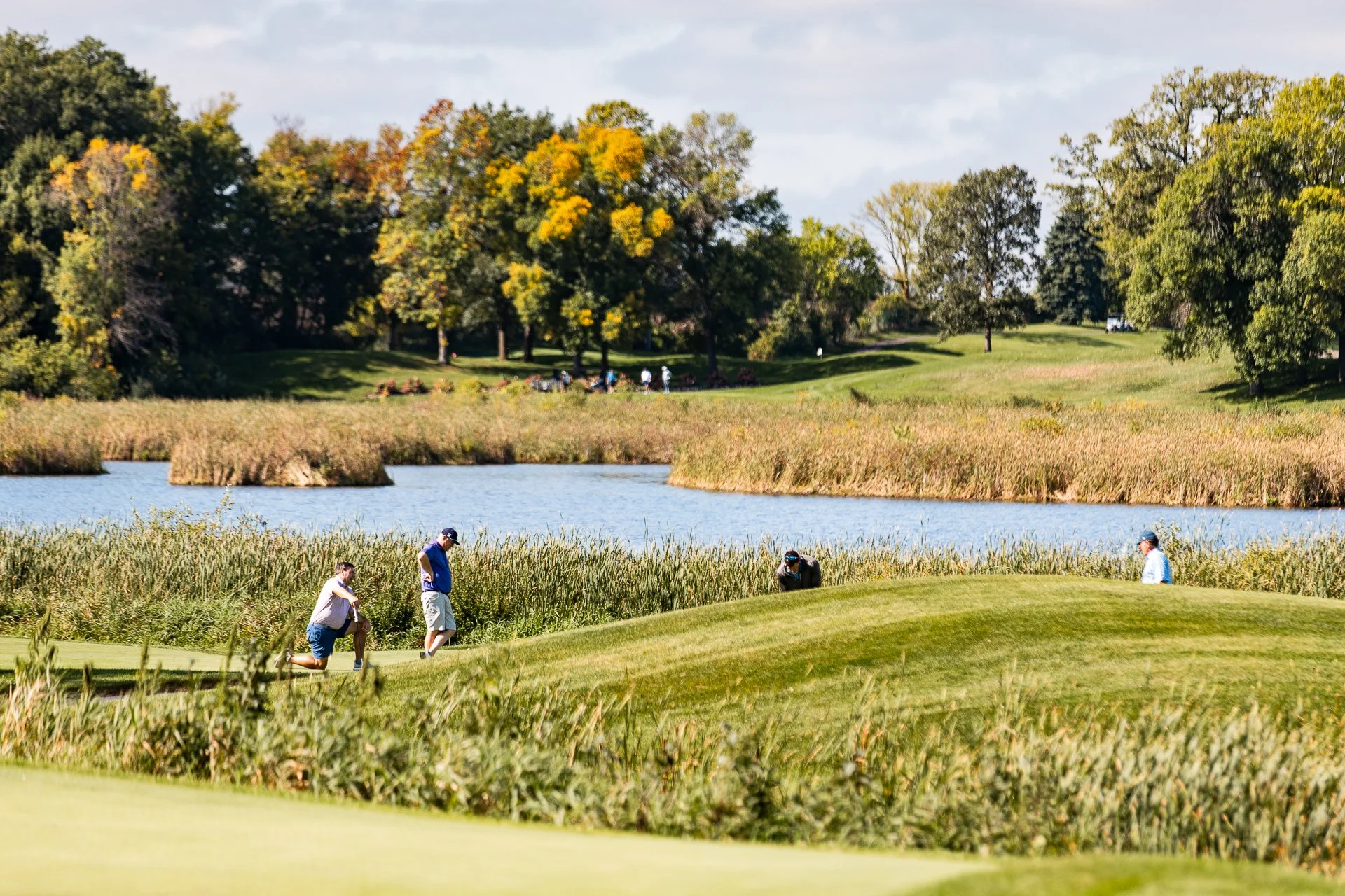 A group of people playing golf near a water body with tall grasses and trees in the background during daytime.