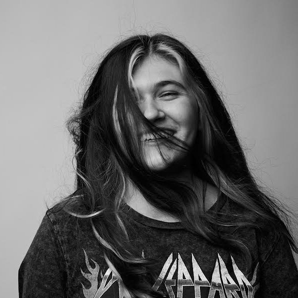 Lilly with long hair smiling with her hair partially covering her face, against a plain background.