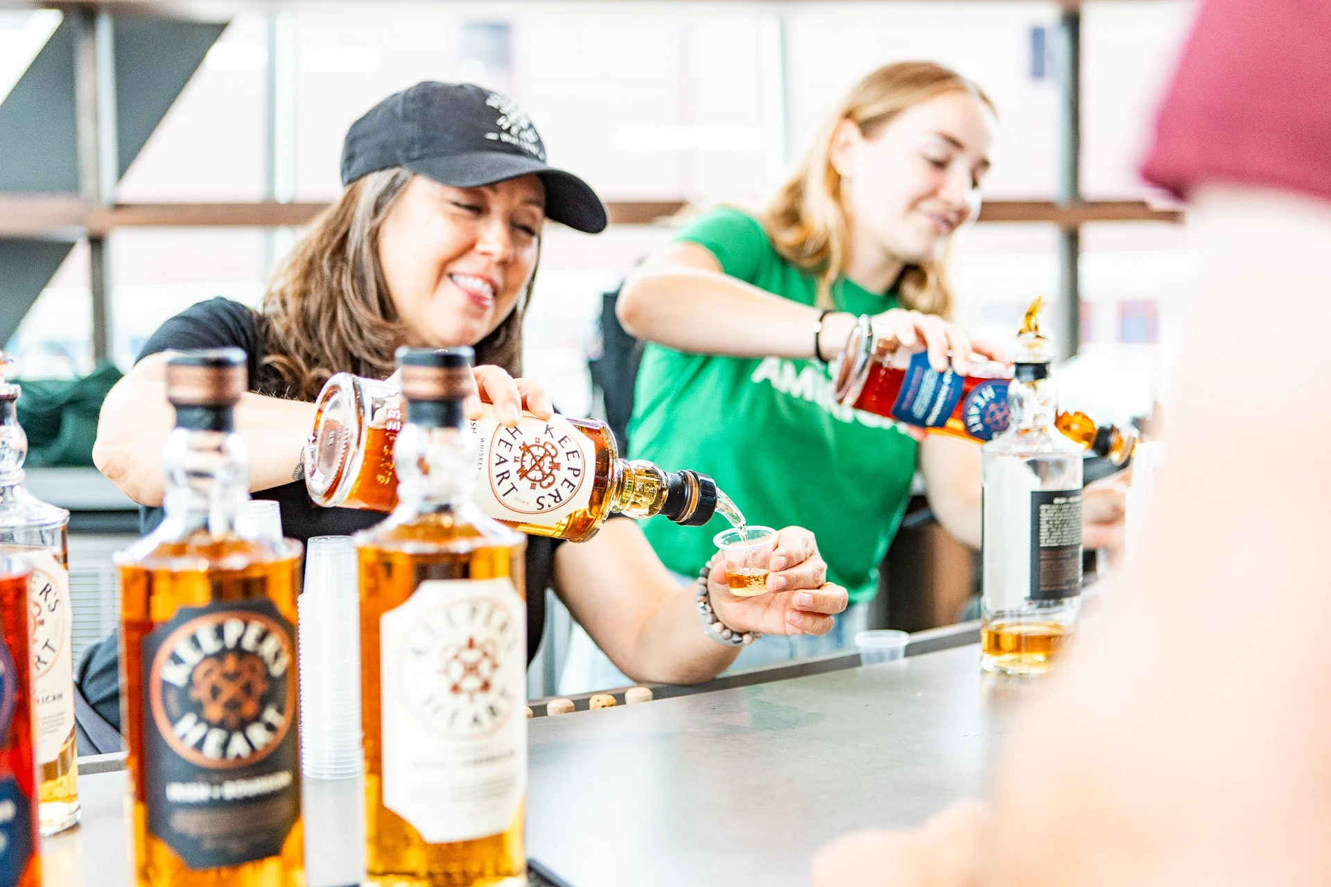 Two women behind a bar counter pouring whiskey into small clear cups, with multiple bottles of whiskey on the counter, in a well-lit modern setting.