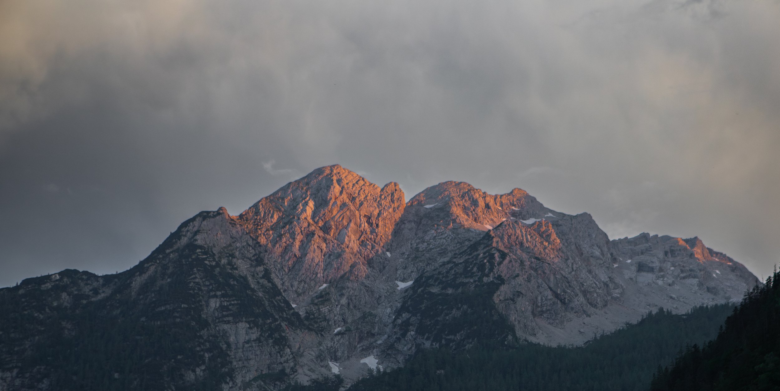 Sunset-lit mountain range with rocky peaks and forested slopes under cloudy sky.