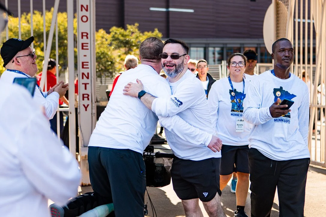 Group of people participating in a charity event, some wearing Minnesota-themed shirts, outside in daylight. Two men are hugging, and others are smiling or engaged, near a gated entrance with a sign that reads 'NO RE-ENTRY,' and trees in the backgrou